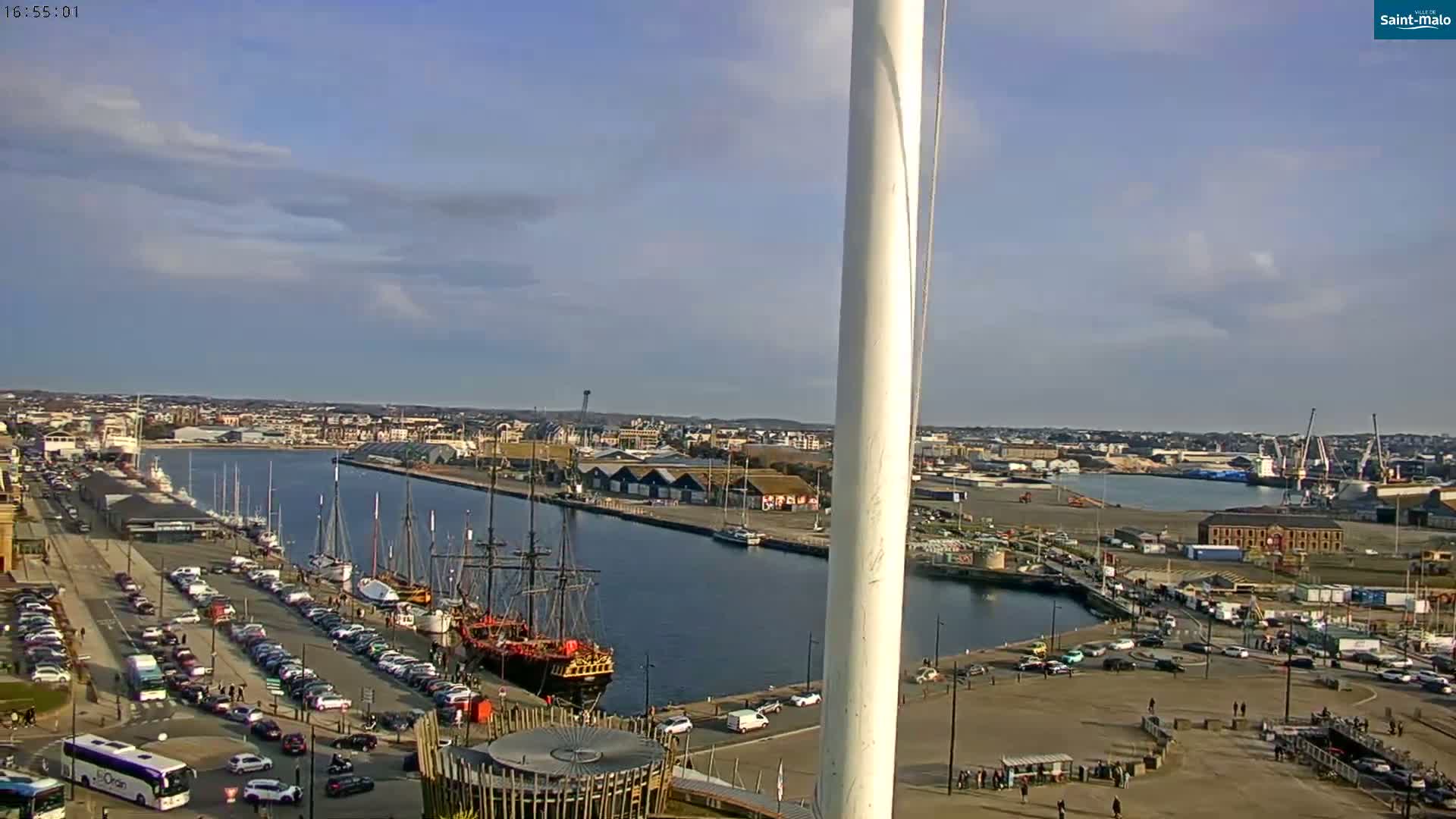 A partly cloudy day overlooks a harbor with numerous docked ships, cars parked along the waterfront, and a city extending into the distance.