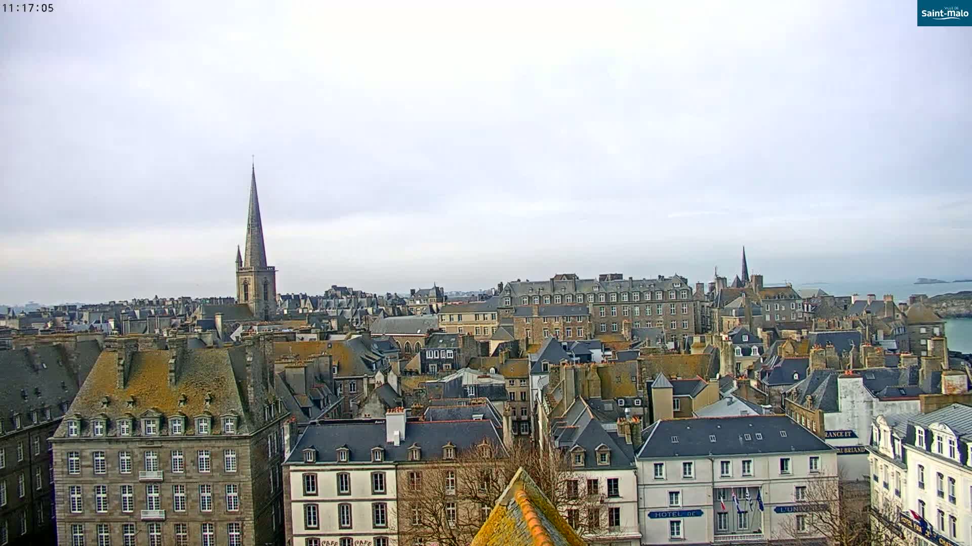A panoramic view of a city with many grey-roofed buildings, a tall church steeple, and a body of water in the distance under an overcast sky.