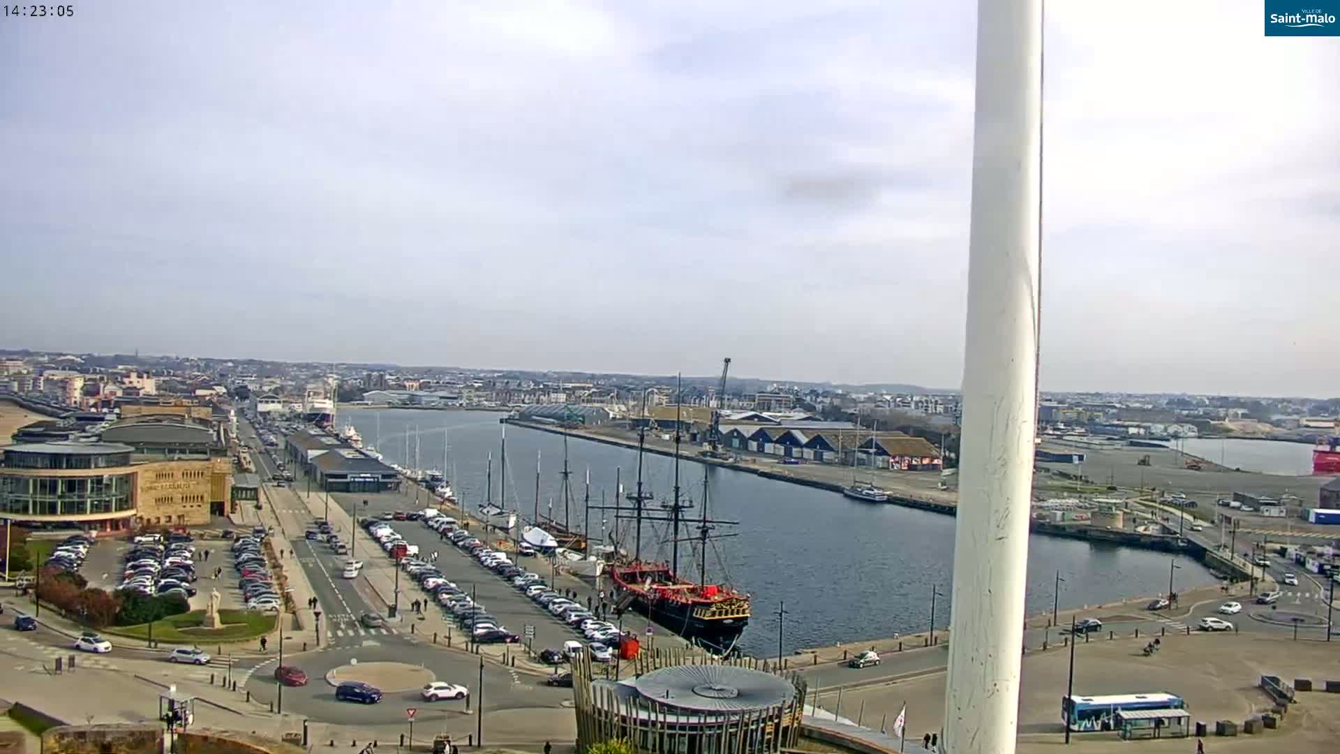 A partly sunny day reveals a harbor scene with a large docked sailing ship, numerous parked cars, and a city stretching into the distance.