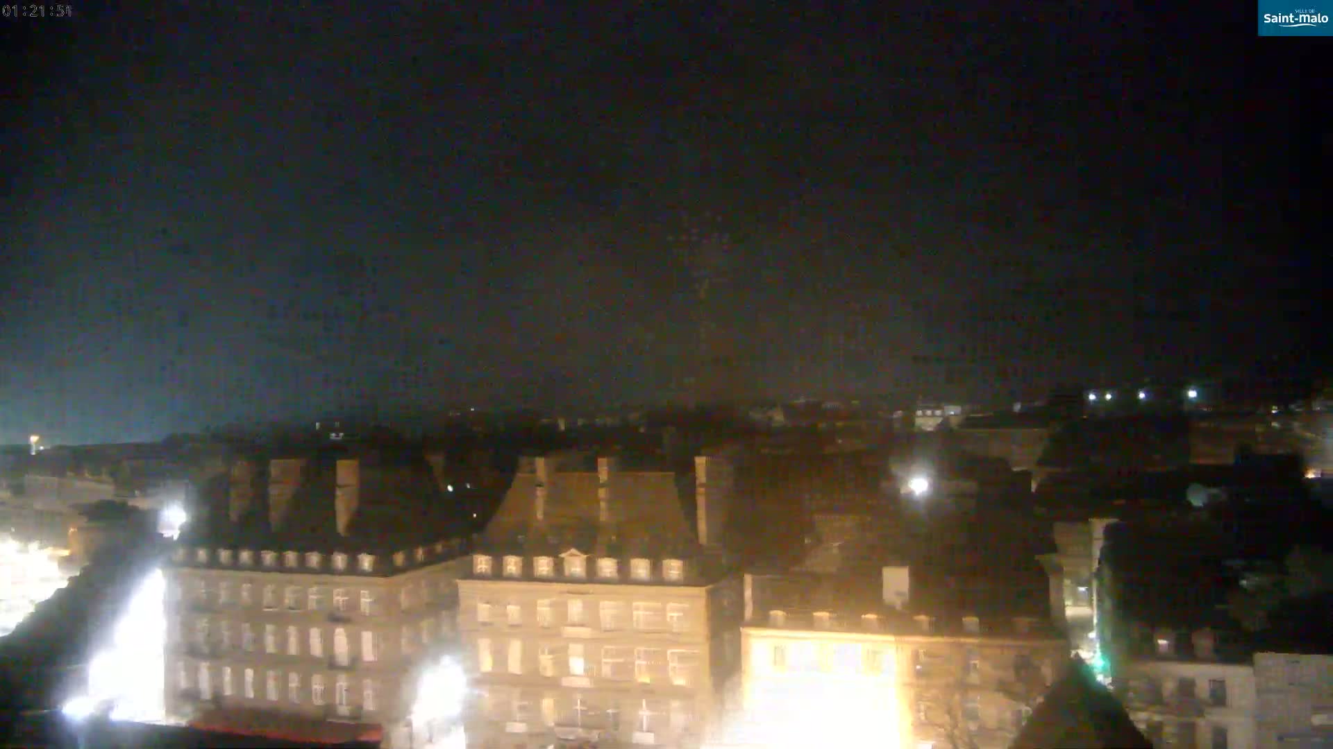 A nighttime, high-angle view shows a city of dark-roofed buildings illuminated by streetlights under a mostly clear, dark sky.