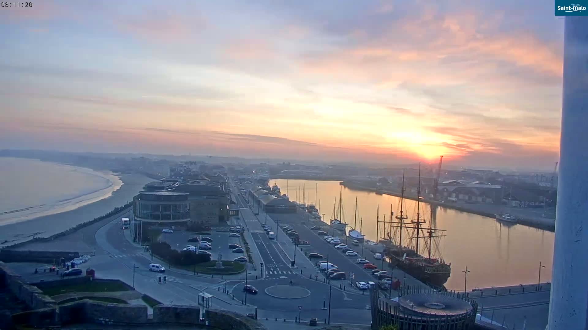 A coastal city at sunrise, with a harbor filled with boats and a sandy beach visible in the distance.