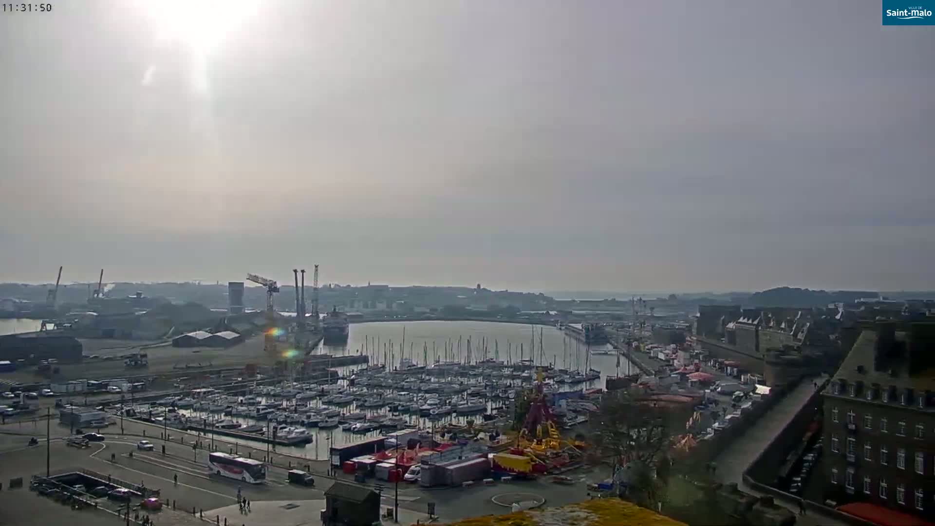 A sunny, hazy day reveals a harbor filled with numerous sailboats, industrial areas, and a portion of a city with buildings along a wall.