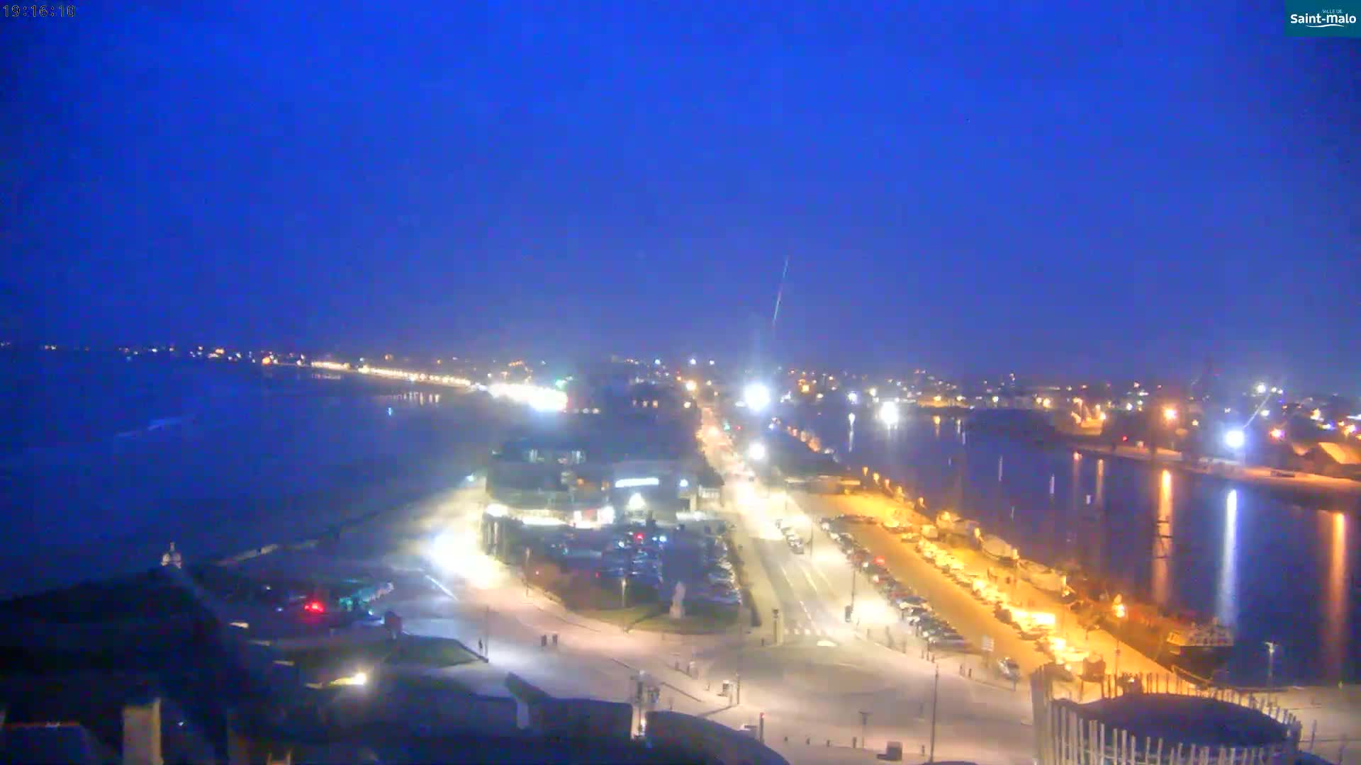 A nighttime view of a coastal town shows a beach, a calm body of water, and illuminated streets and buildings.