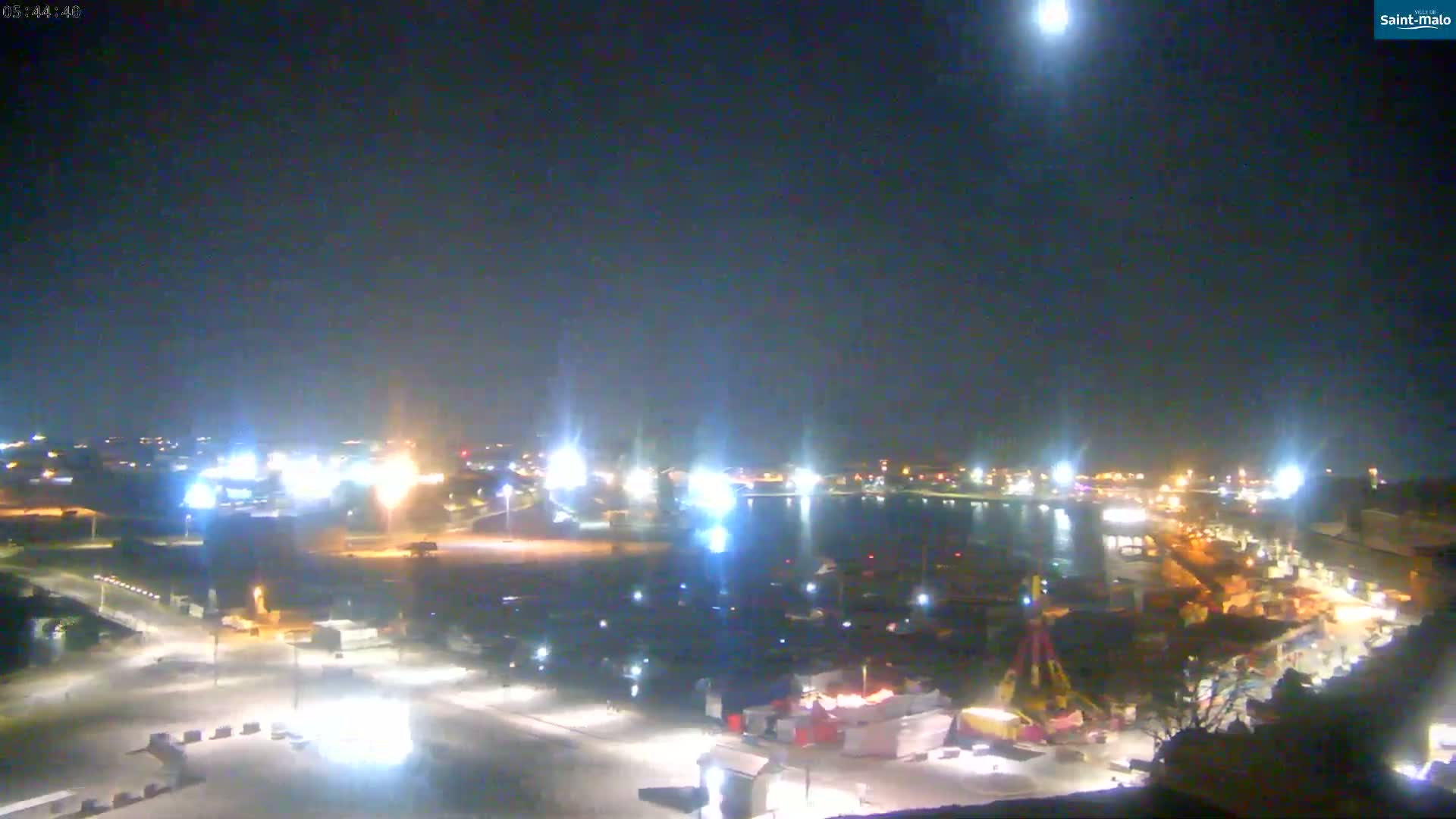 A nighttime, high-angle view of a harbor and city area with bright lights illuminating the scene under a clear, dark sky.