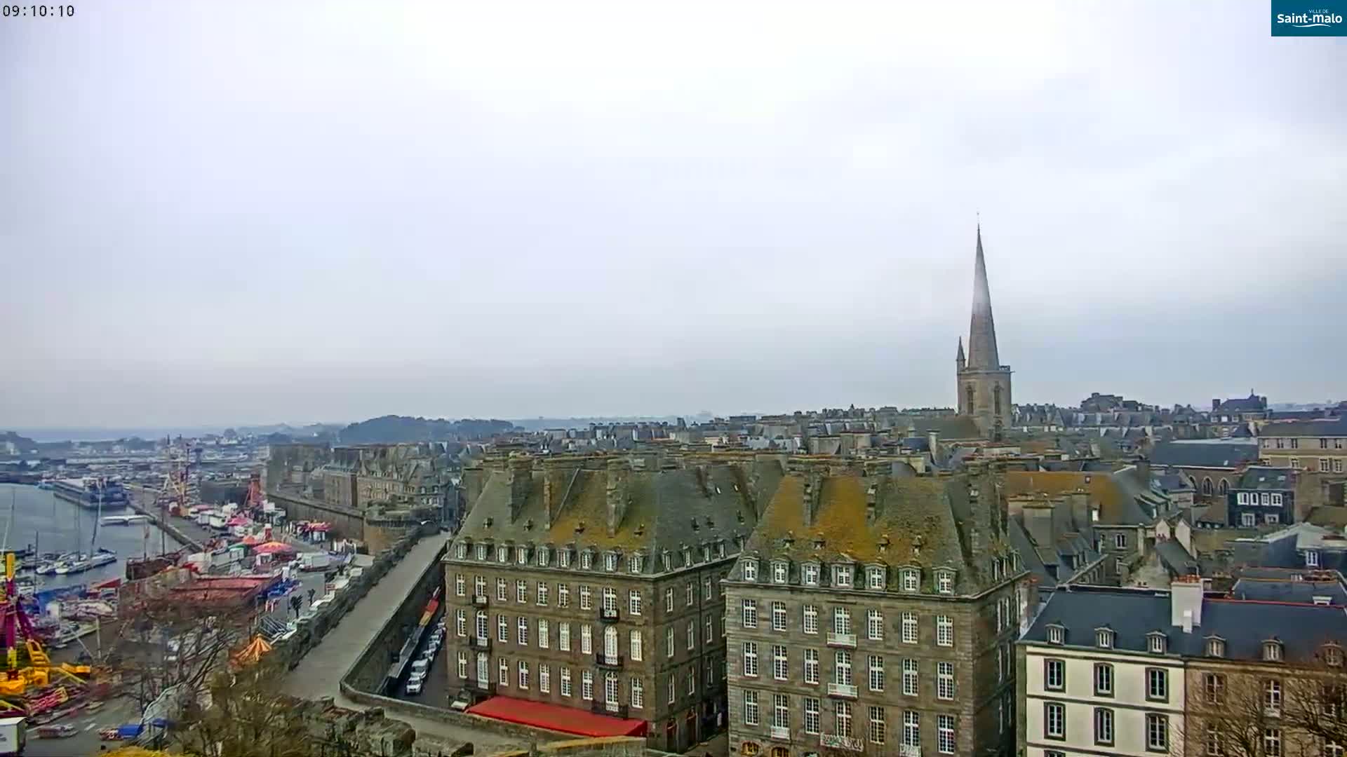A high-angle, long shot of a European city on an overcast day, showing a harbor with boats, a walled area with buildings, and a tall church spire in the distance.