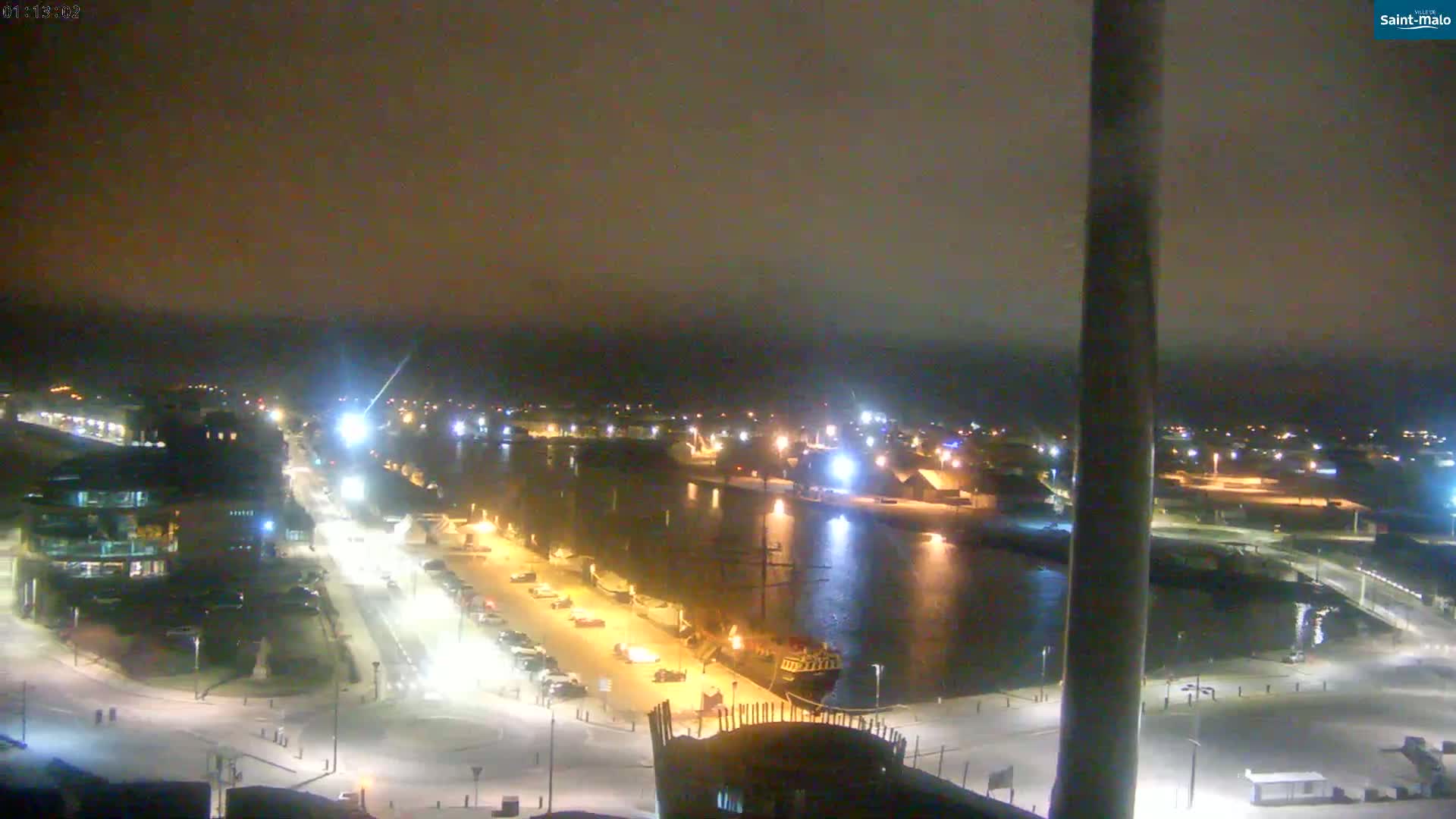 A nighttime view of a snow-covered harbor town with a calm body of water, illuminated buildings and parked cars along the waterfront.