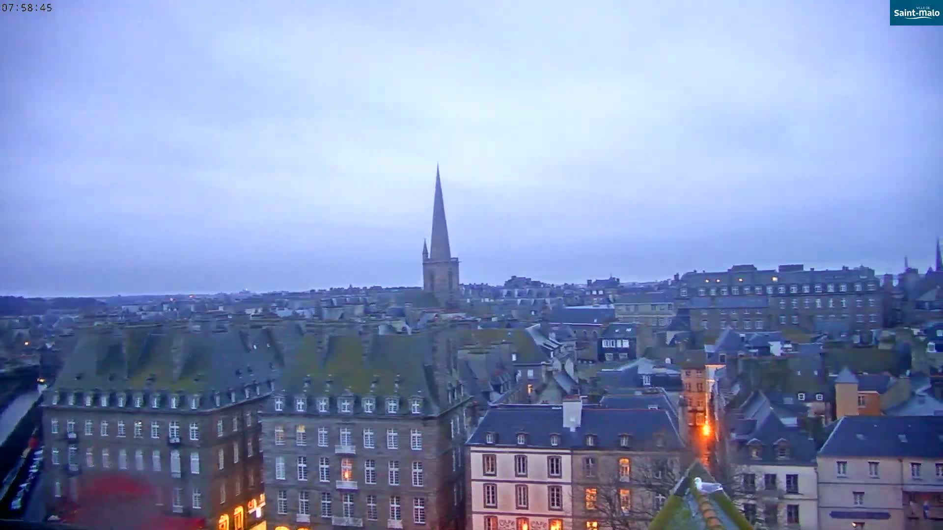 A high-angle, long shot shows a city of many grey-roofed buildings under a pale, overcast sky at dusk.