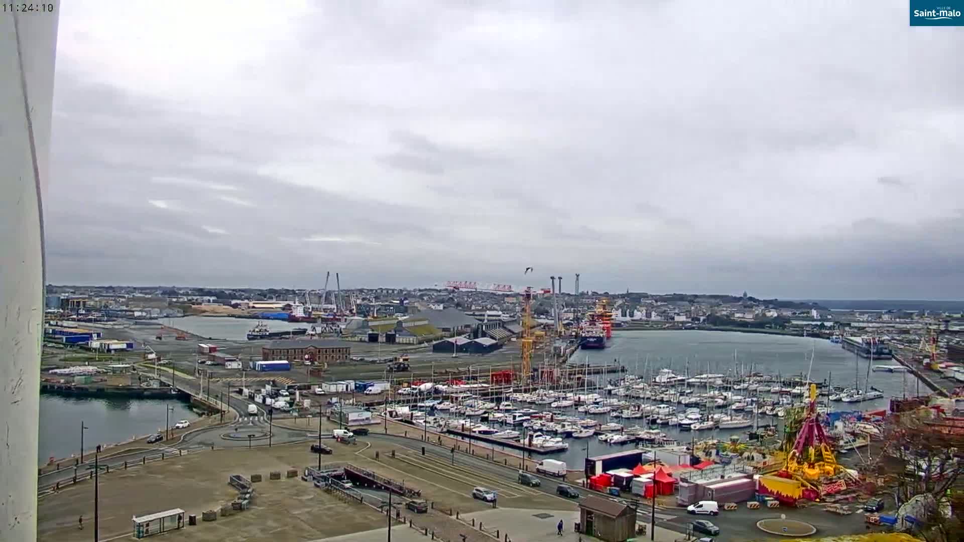 An overcast day shows a harbor filled with numerous boats and ships, industrial areas, and a large amusement park ride near a roadway.