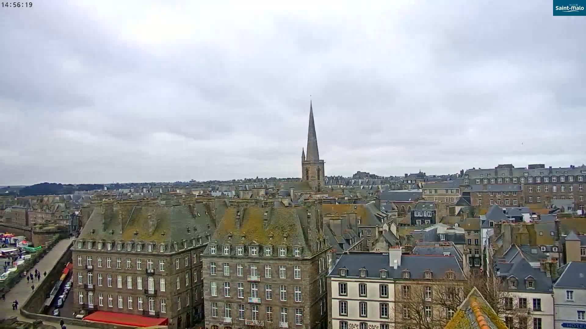 A panoramic view of a European city under an overcast sky shows many stone buildings with steeply pitched roofs, and a tall church spire in the distance.