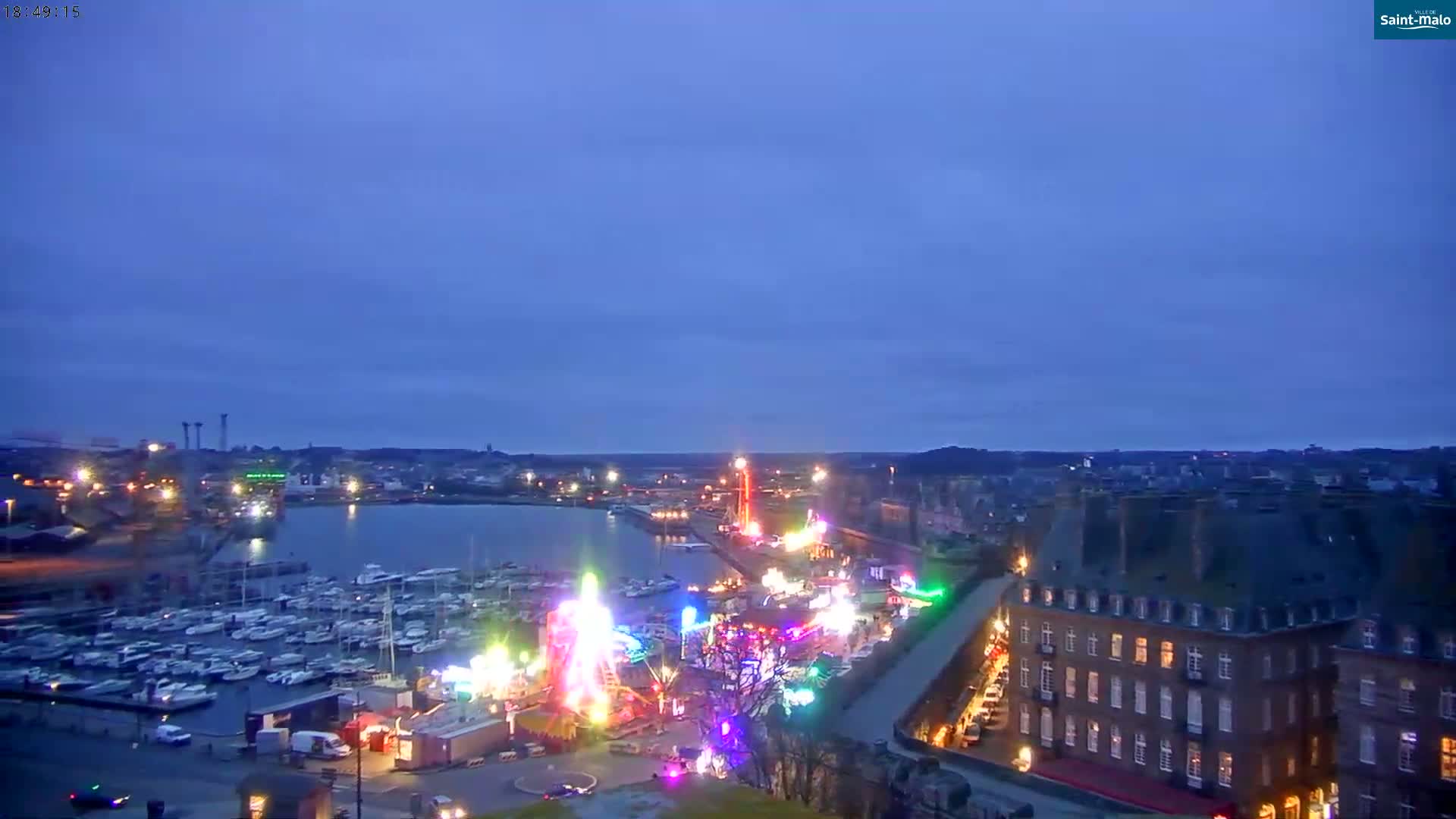 A nighttime, twilight view of a harbor filled with boats, a brightly lit amusement park, and a large building.