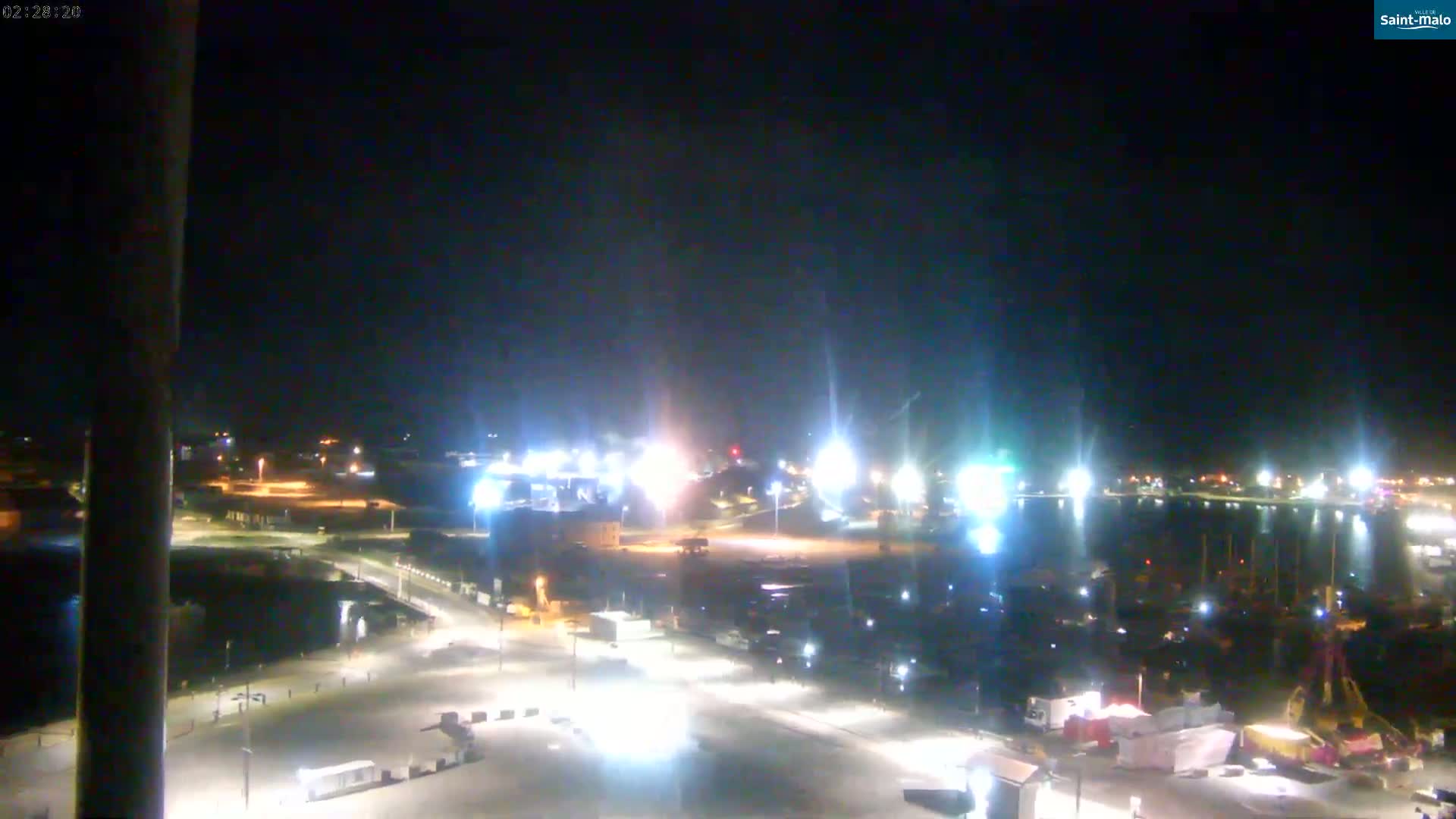 A nighttime, brightly lit harbor scene shows a port area with docked boats and vehicles in a parking lot under a clear, dark sky.