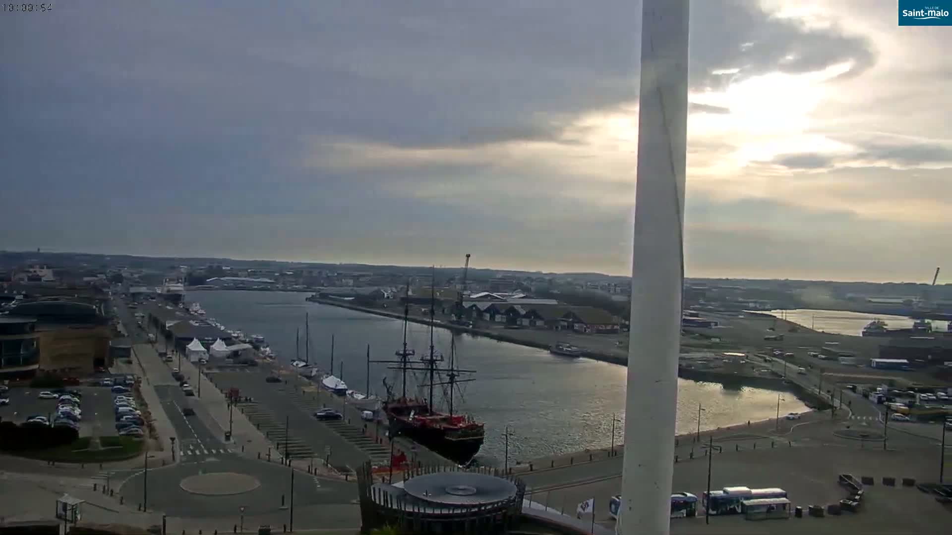 An elevated view of a harbor shows a large sailing ship docked near a city under partly cloudy skies.