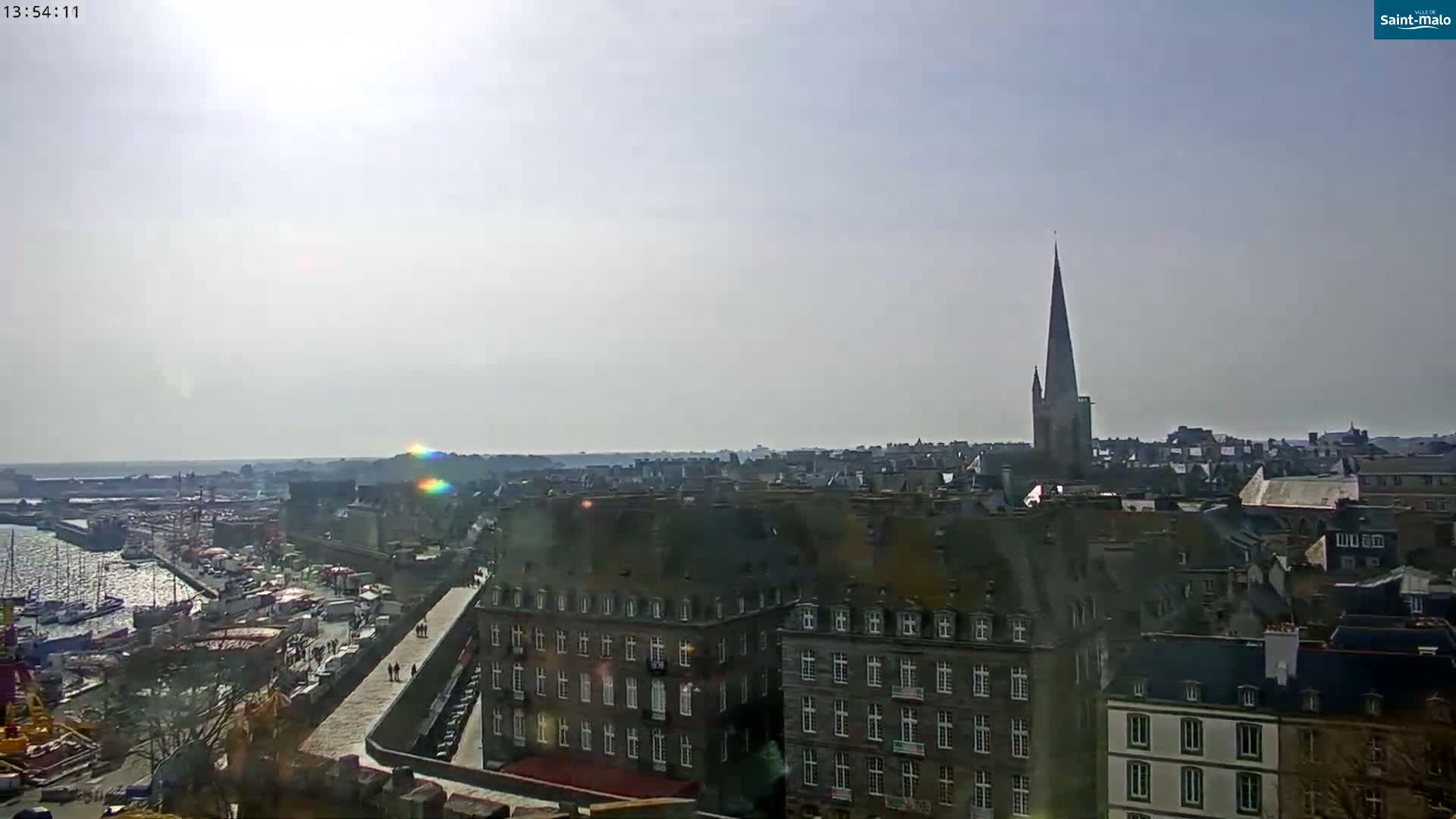 A sunny day reveals a panoramic view of a coastal city with a harbor, featuring a tall church spire and numerous buildings of similar architectural style.