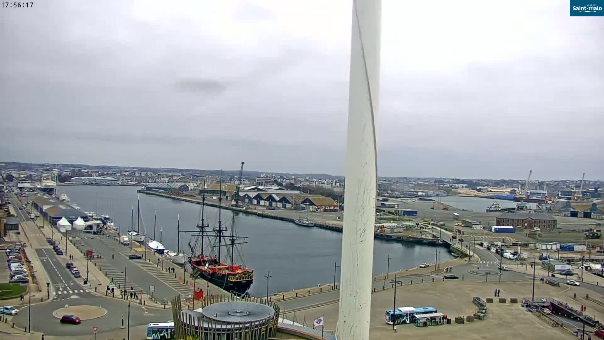 An overcast day reveals a harbor scene with a large sailing ship docked near a waterfront city, featuring a tall, slender white structure in the foreground.