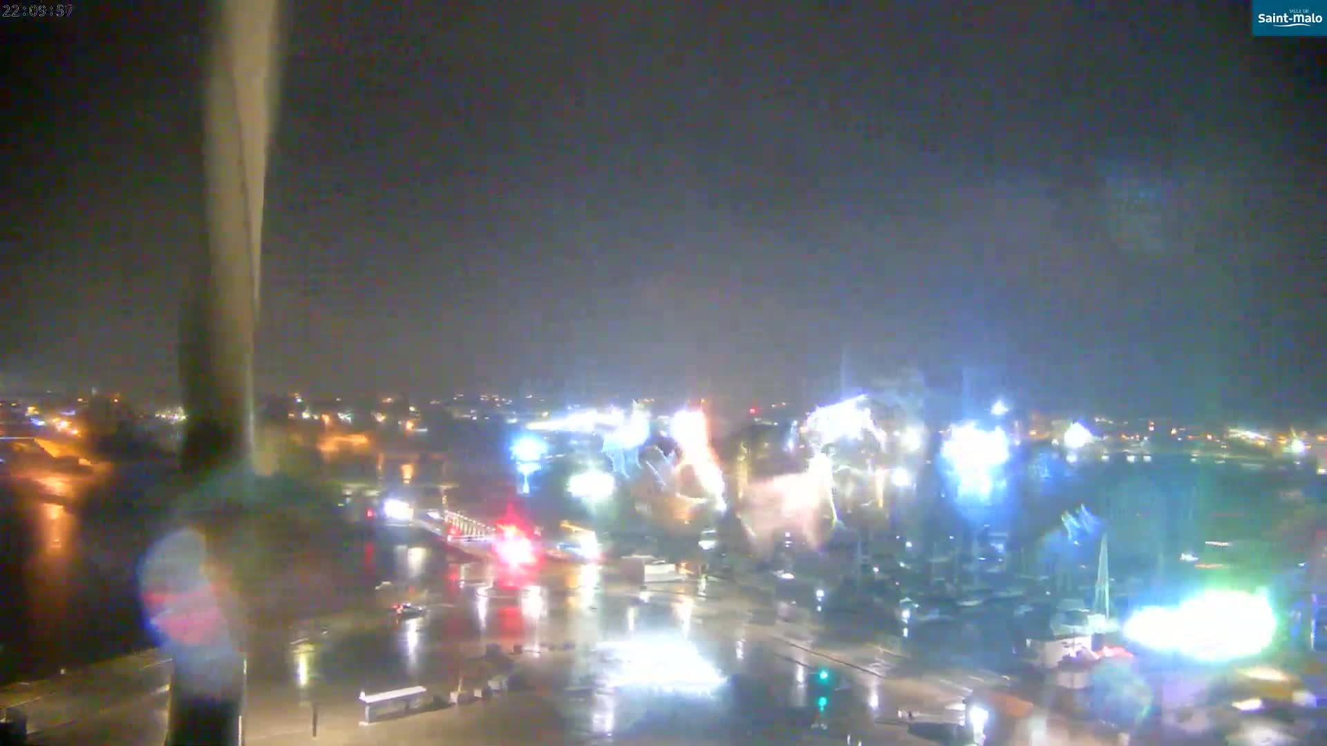 A nighttime view of fireworks exploding over a rain-soaked harbor, with city lights reflecting on wet streets and a tall structure visible in the foreground.