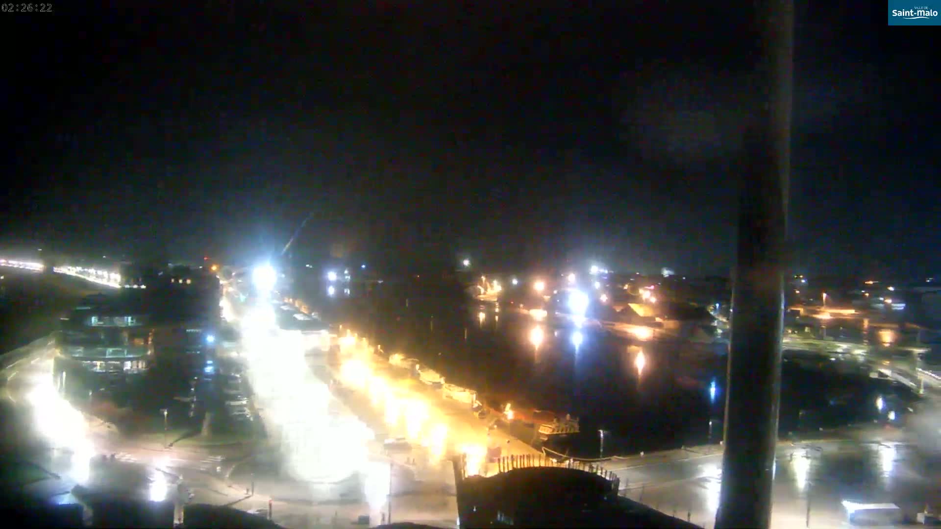A nighttime aerial view shows a harbor with boats and buildings illuminated by streetlights and other lights, under a dark, clear sky.