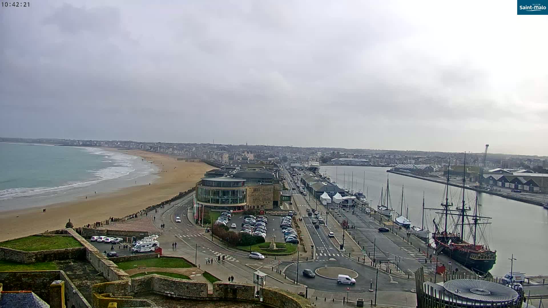 An expansive view of a coastal city on an overcast day, showcasing a sandy beach, a harbor filled with boats, and a large sailing ship docked near a stone building.