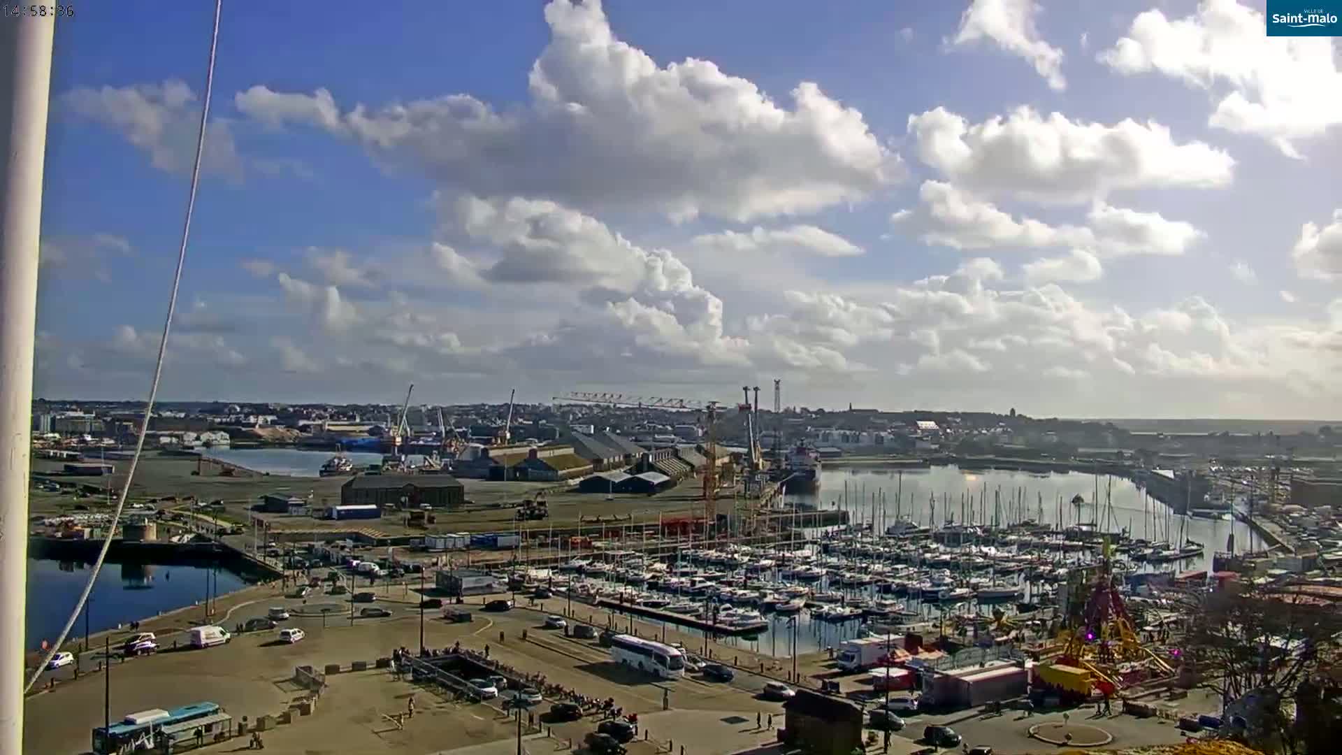 A partly cloudy day reveals a harbor filled with numerous sailboats and other vessels, surrounded by a town and industrial areas.