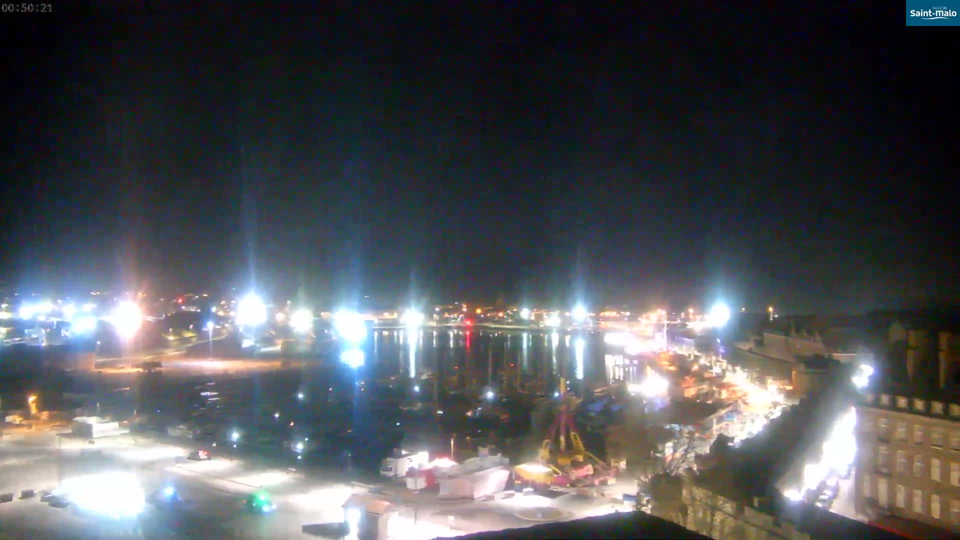 A nighttime, brightly lit harbor scene with boats, buildings, and streets visible under a clear, dark sky.