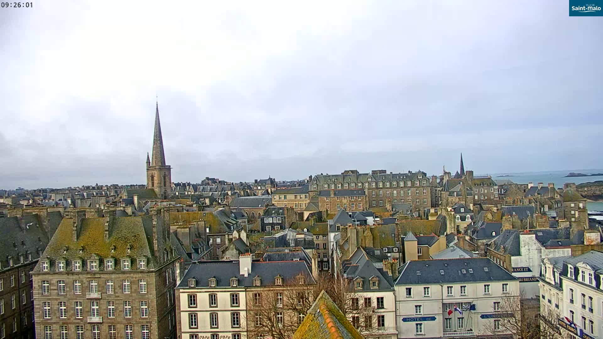 A panoramic view of a city with many closely-packed buildings, a prominent church spire, and a calm body of water visible in the distance under an overcast sky.
