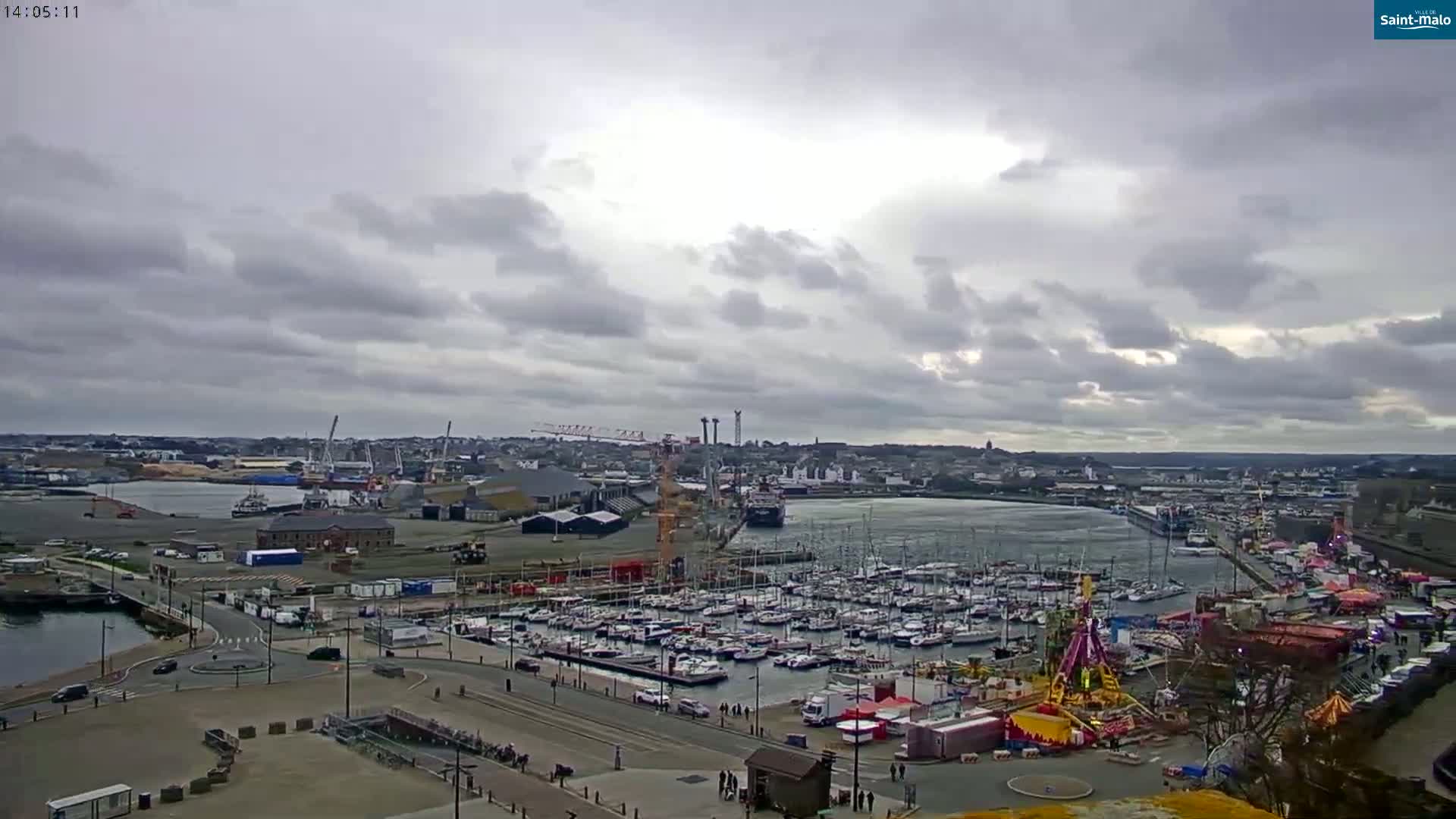 A harbor filled with numerous sailboats and a fairground is visible under an overcast sky.
