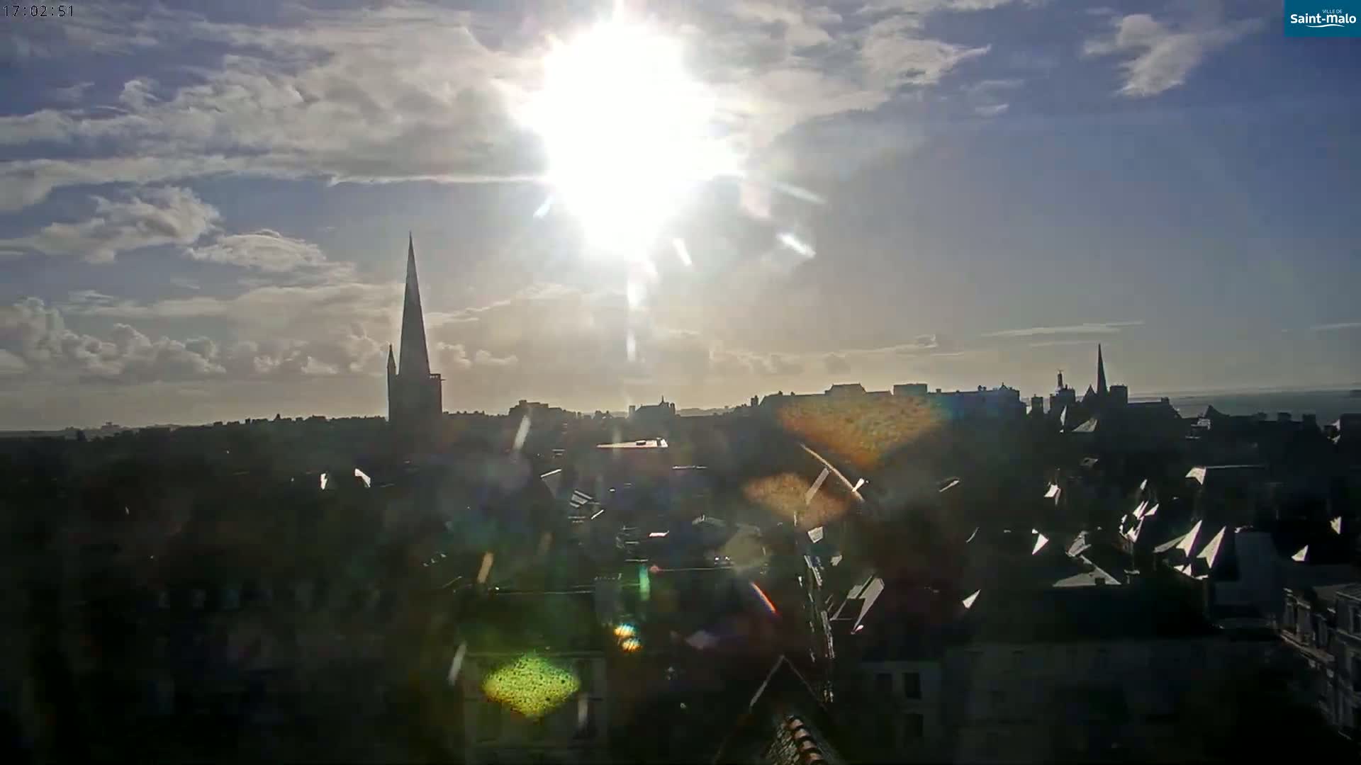 A partly cloudy daytime scene shows a city skyline with a prominent church spire, the sun shining brightly in the sky.