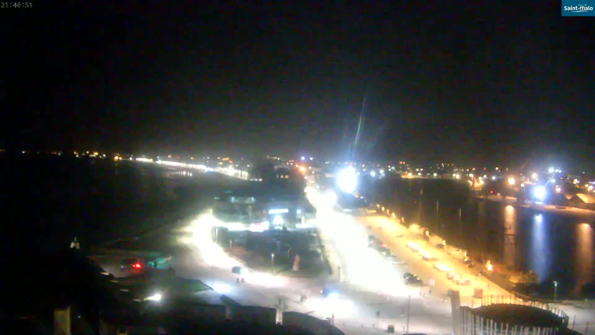 A nighttime, high-angle view shows a snow-covered road with traffic and nearby buildings illuminated by lights, beside a dark body of water.