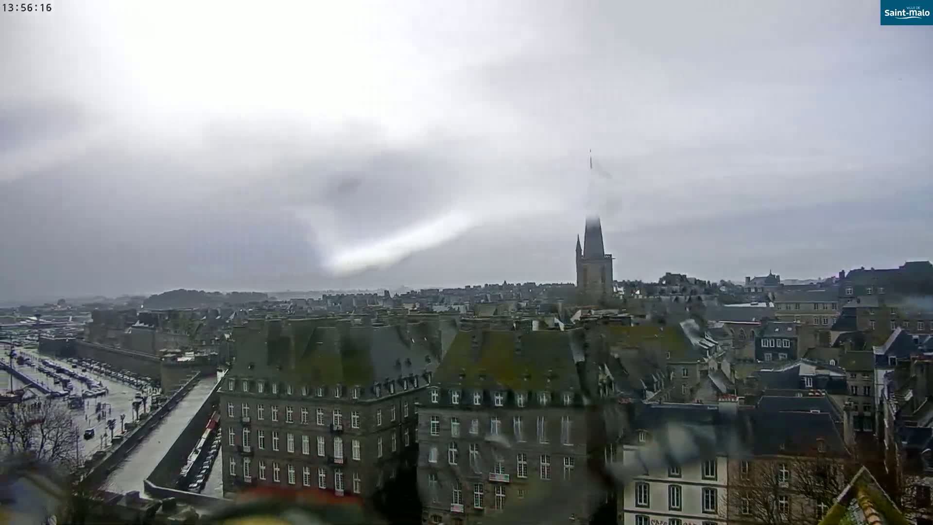 A rain-streaked view shows a European city with a tall church spire and many grey buildings under an overcast sky.