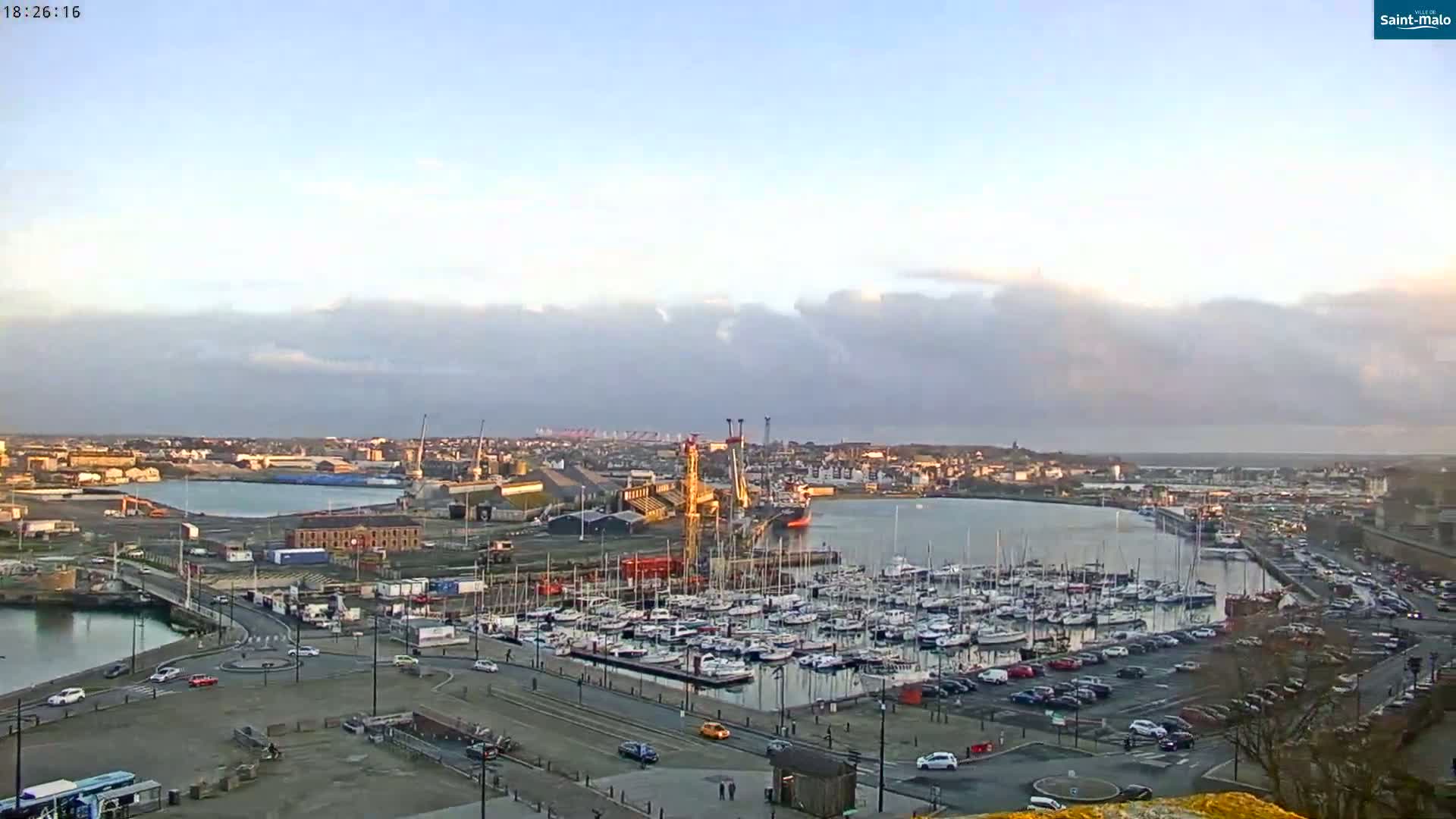 A harbor filled with numerous sailboats and a large cargo ship is shown under a partly cloudy sky.