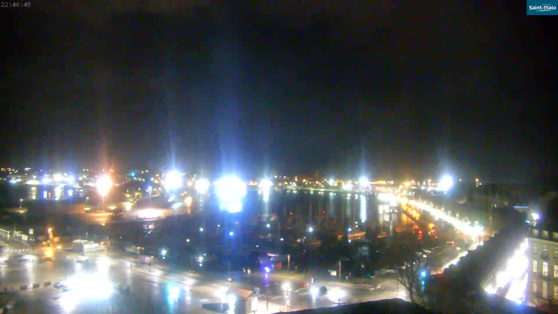 A nighttime, wet view of a harbor town with brightly lit streets and docked boats, under a dark sky.