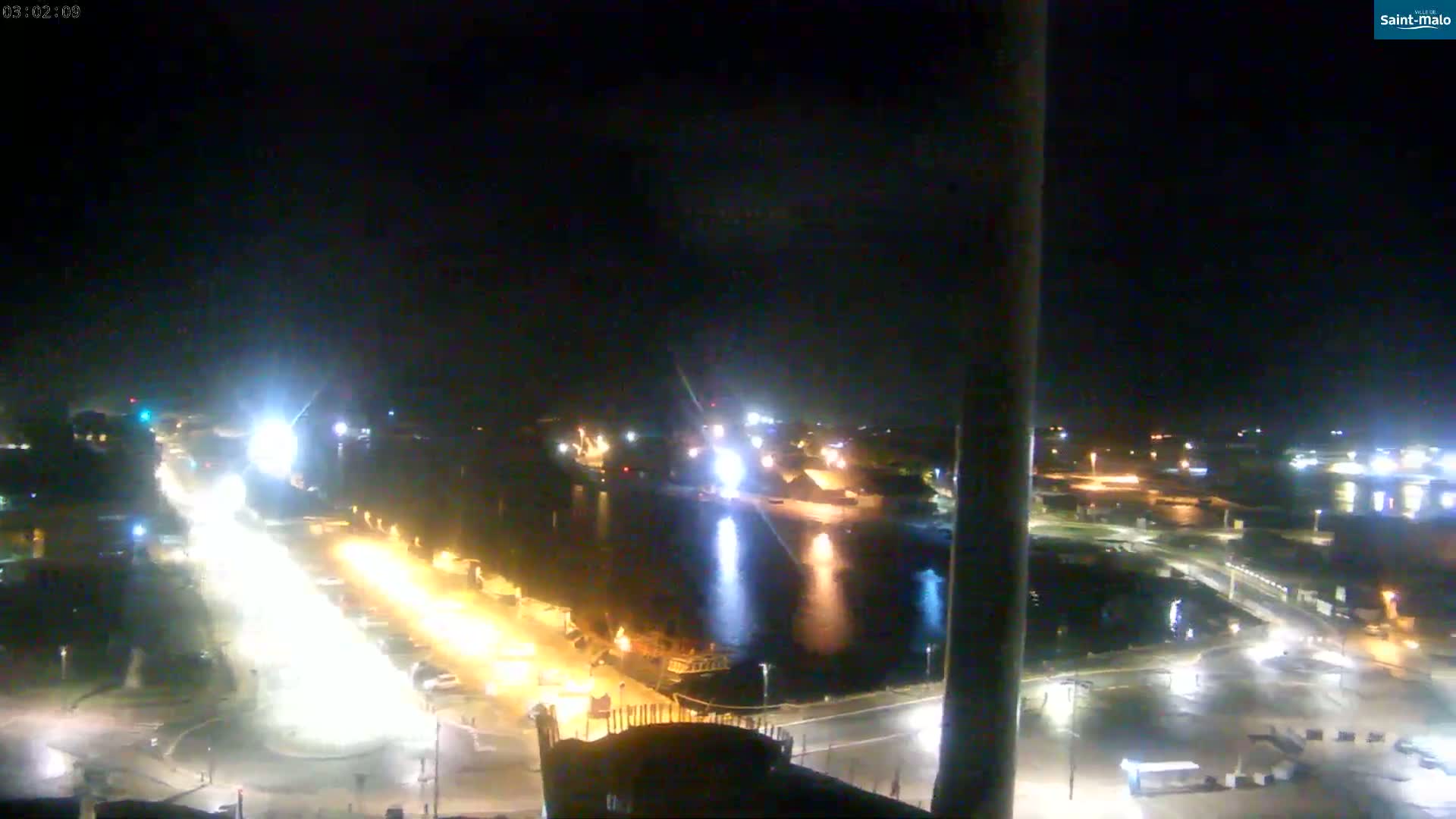 A nighttime, high-angle view of a harbor with brightly lit roads and docked boats, under a dark sky.