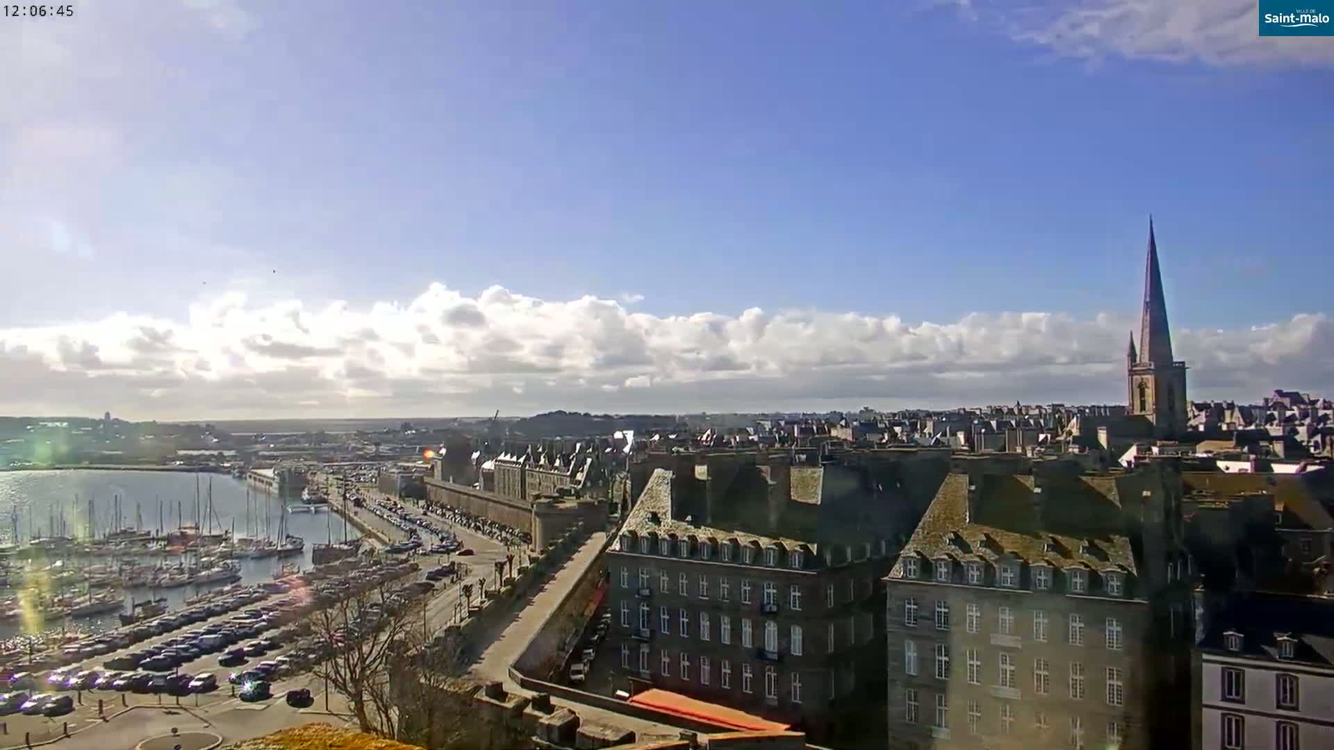 A partly cloudy day reveals a harbor filled with sailboats and cars parked along a waterfront city with a tall church spire in the background.