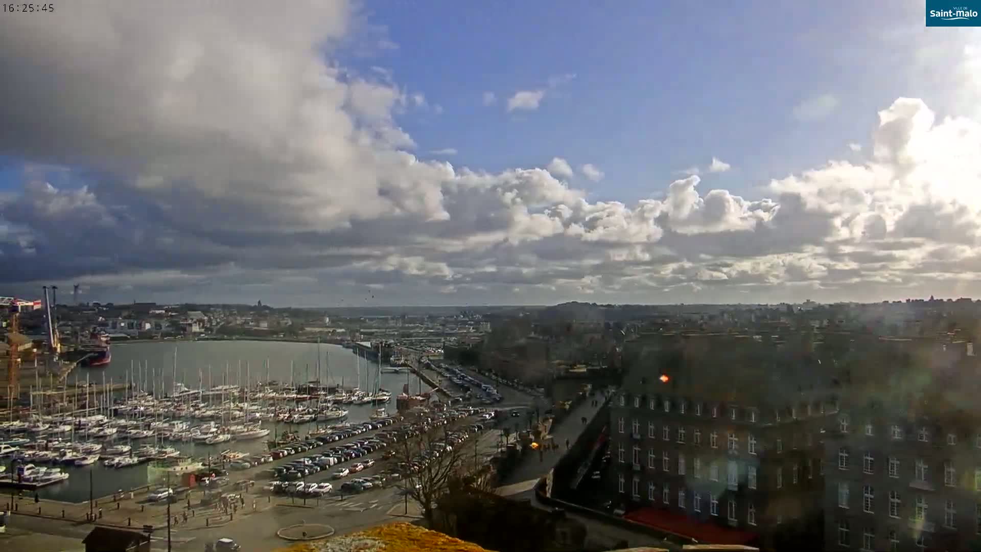 A partly cloudy sky overlooks a harbor filled with sailboats and a city with parked cars and buildings.
