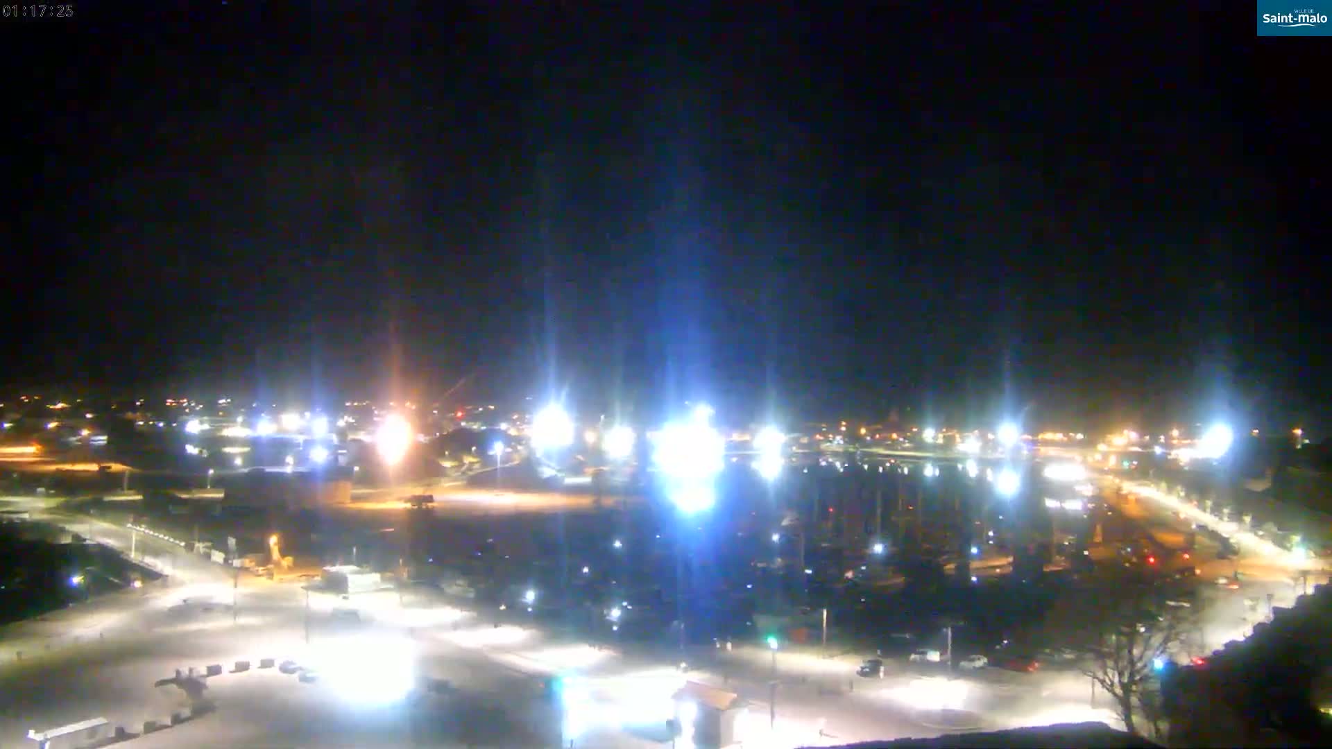 A nighttime, brightly lit harbor scene with numerous boats and vehicles, under a clear, dark sky.