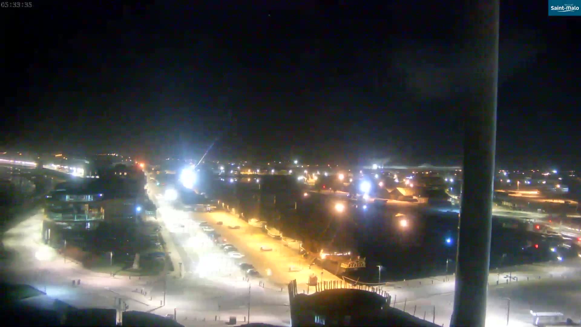 A nighttime, snow-covered harbor scene shows a parking lot, buildings, and boats illuminated by streetlights.