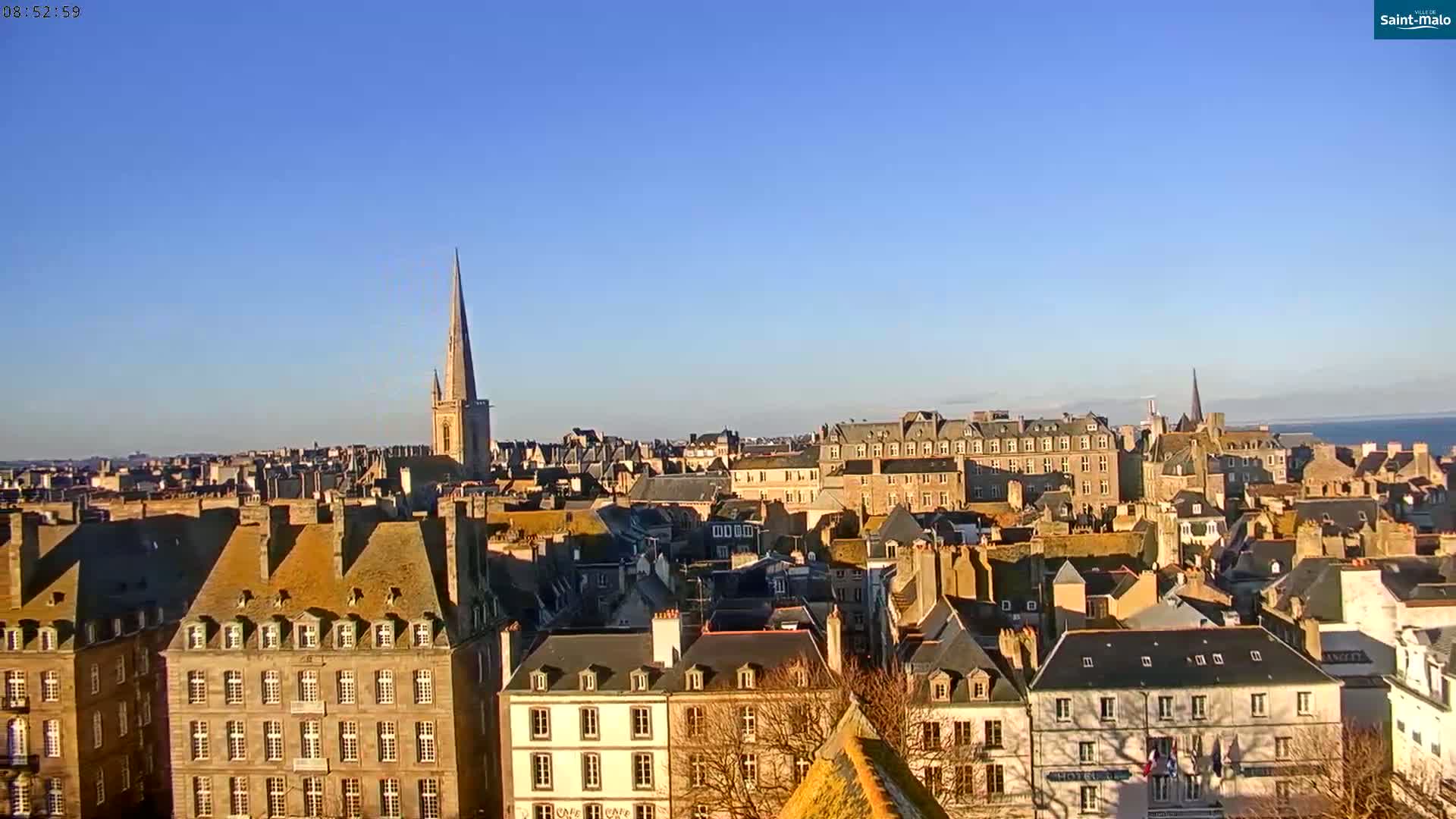 A panoramic view of a European city on a clear, sunny day, showing many stone buildings with dark roofs, and a tall church spire in the distance.