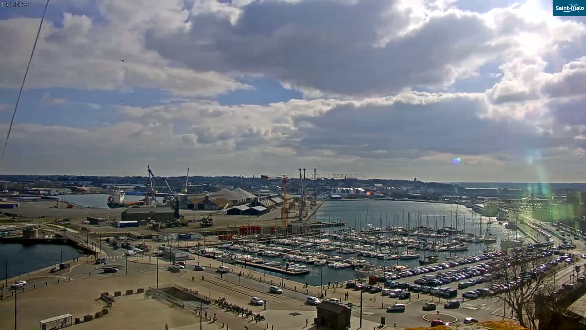 A partly cloudy day overlooks a harbor filled with numerous sailboats and a large parking lot, with cranes and industrial buildings visible in the background.