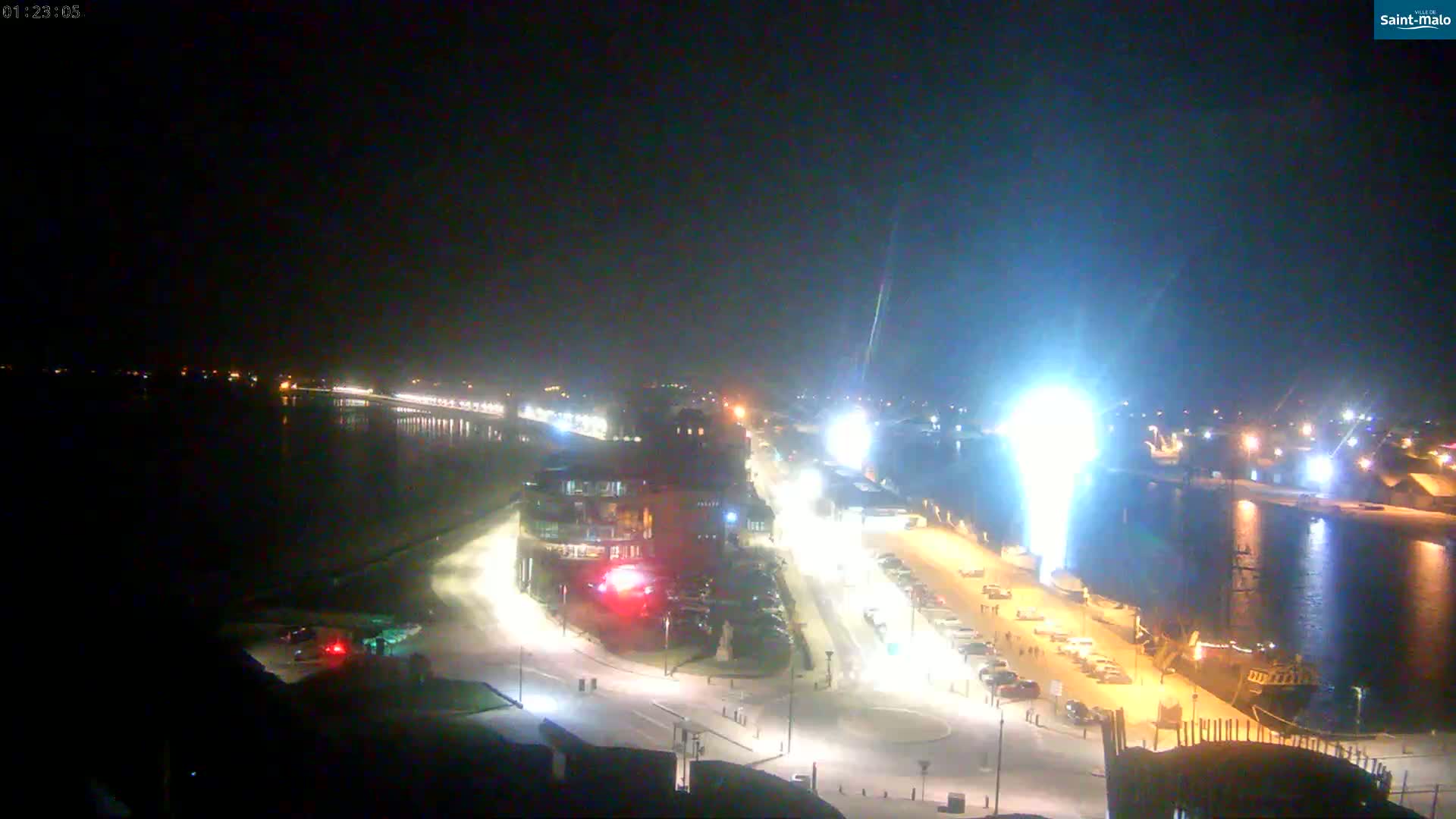 A nighttime aerial view of a waterfront area shows a body of water, a roadway with parked cars, buildings with lights, and a large ship at the dock, under clear skies.