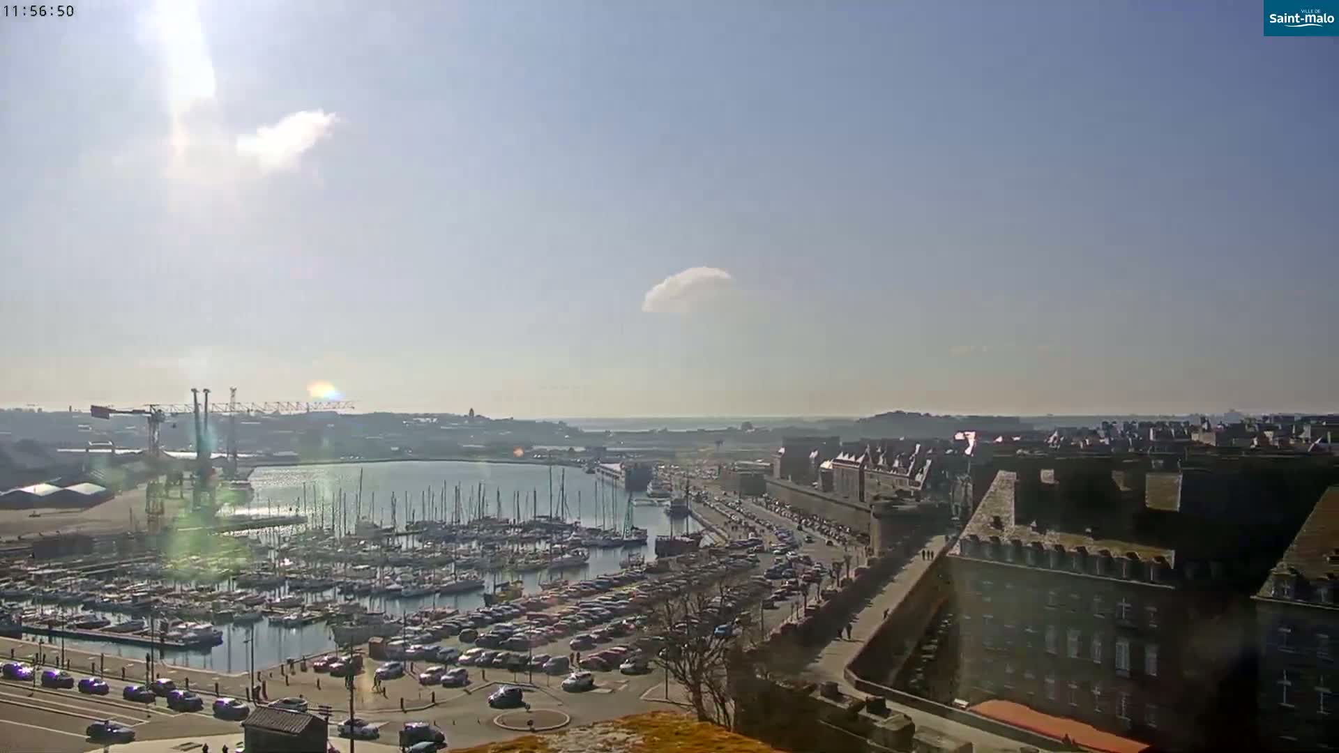 A sunny day reveals a harbor filled with sailboats and a parking lot next to a walled city.