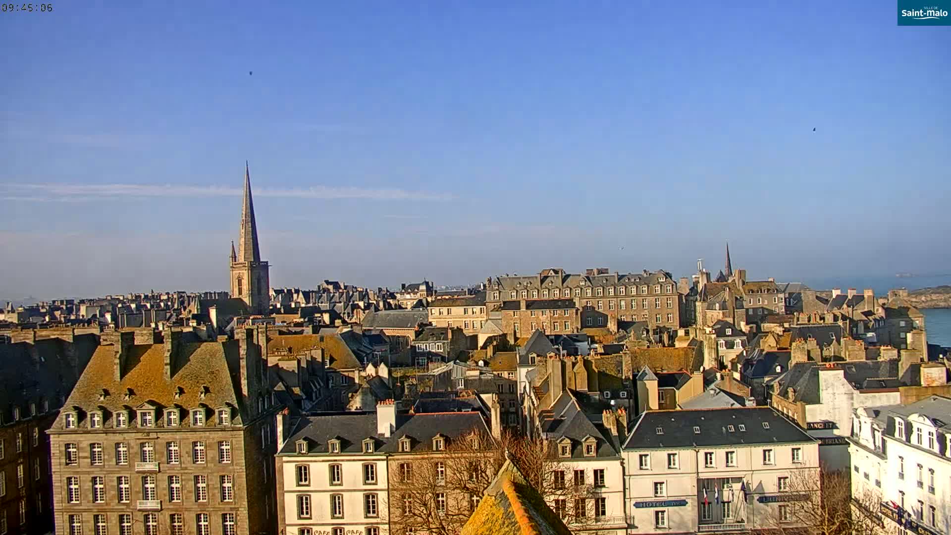 A sprawling cityscape of closely packed buildings with steeply pitched roofs under a clear blue sky, overlooking a calm body of water in the distance.