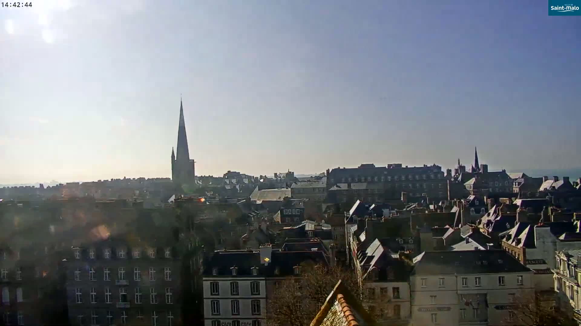 A sunny, hazy day reveals a panoramic view of a European city with numerous rooftops and a tall spire in the distance.
