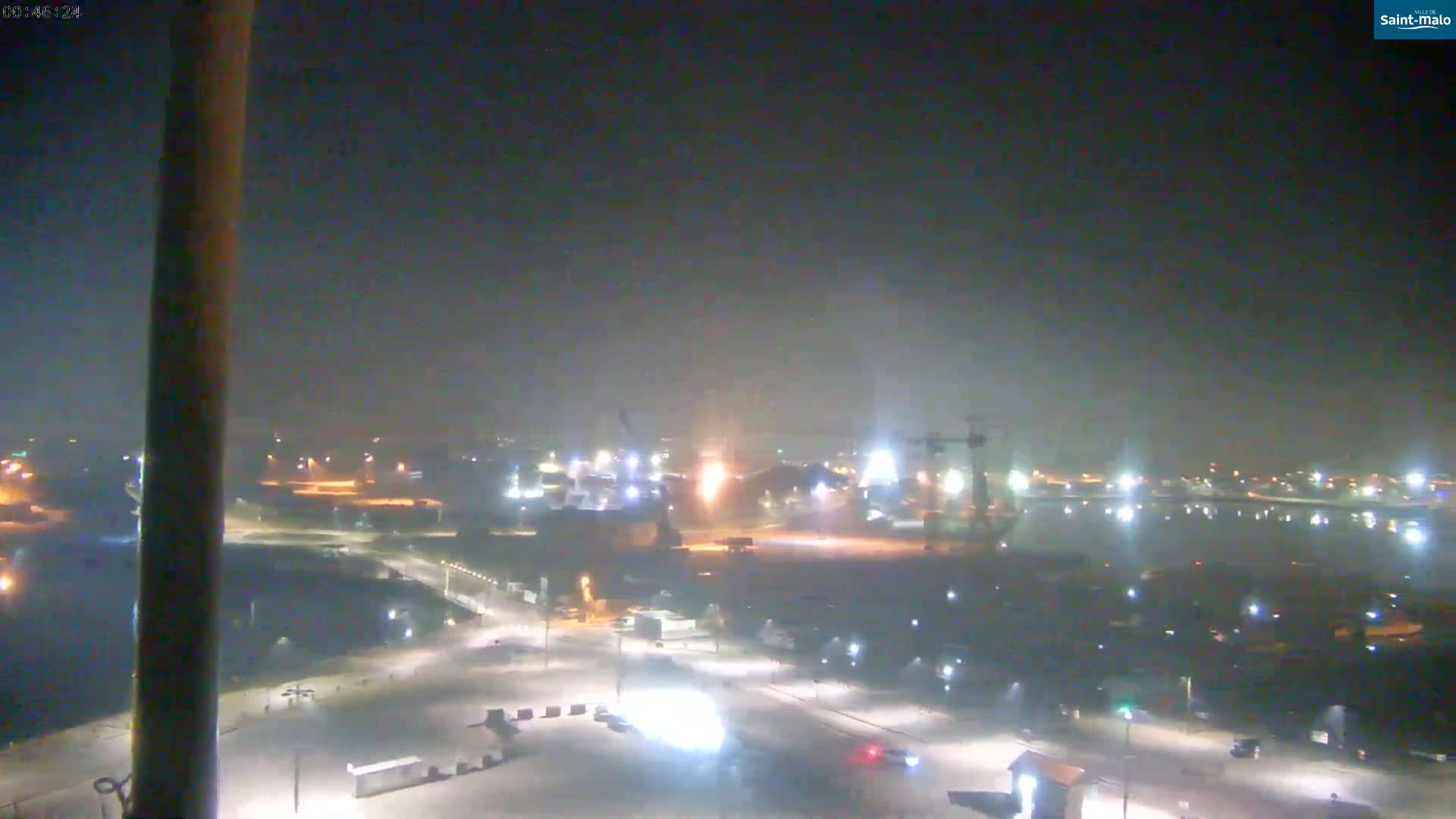 A nighttime, snowy scene shows a harbor area with industrial buildings and cranes illuminated under a hazy sky.
