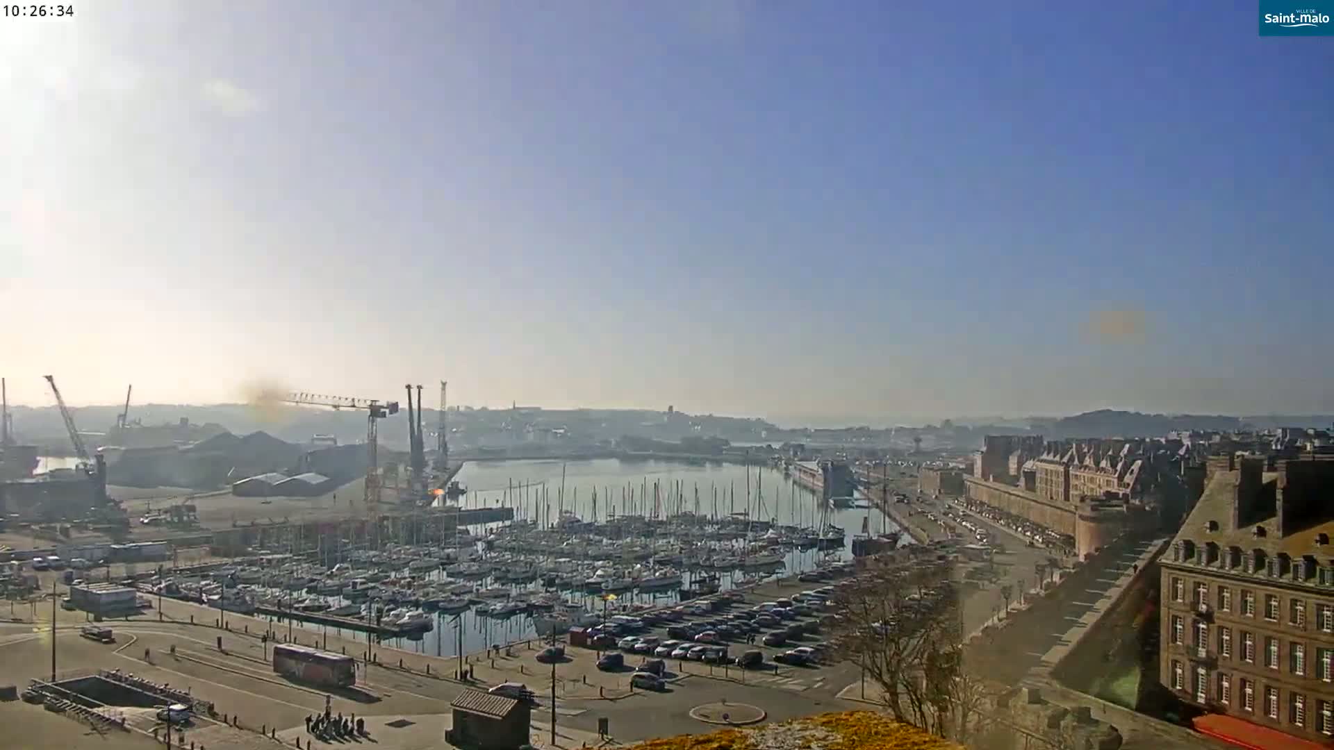 A sunny harbor scene shows a marina filled with sailboats, a parking lot, and a city with historic-looking buildings under a clear sky.
