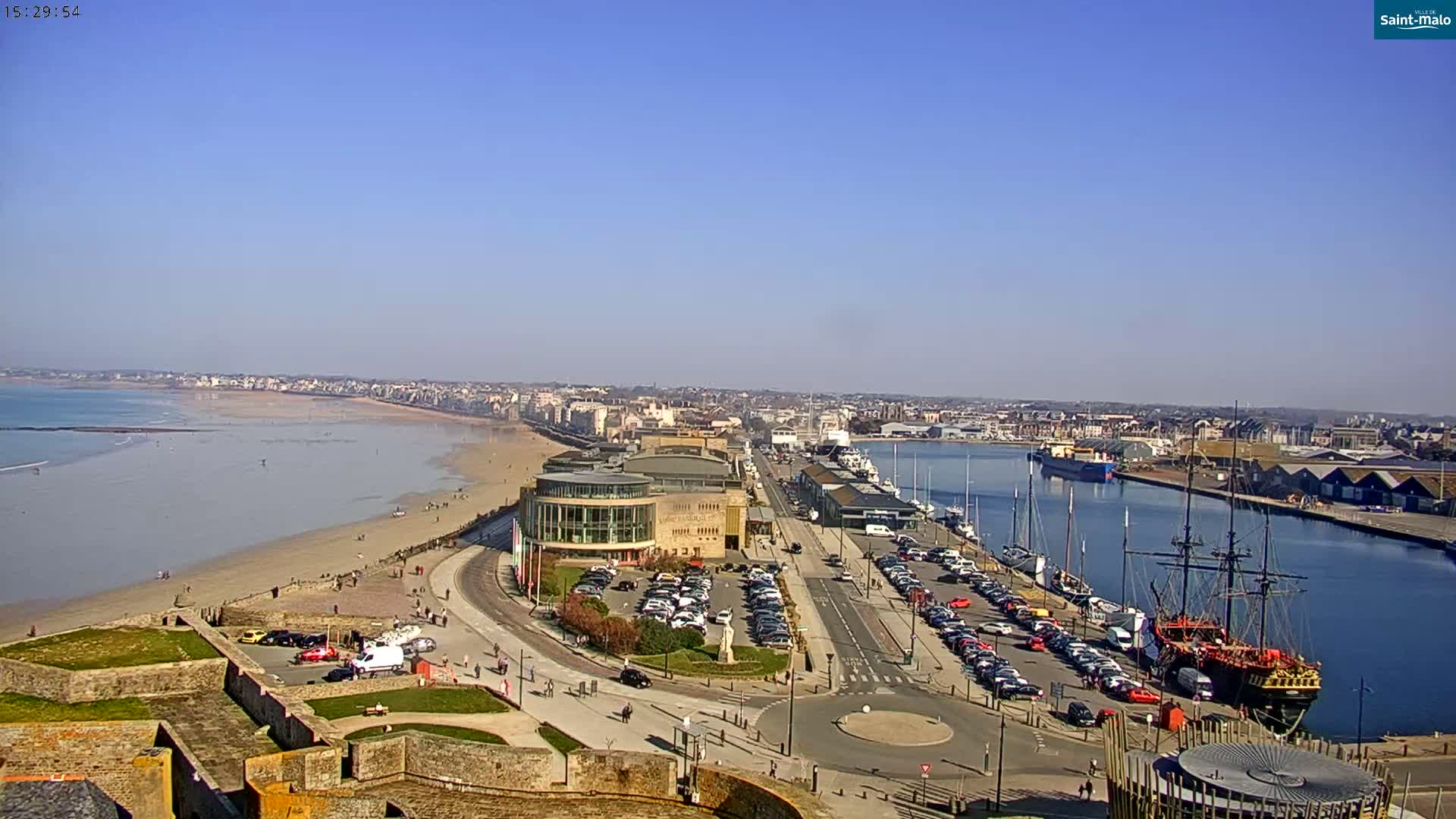 A wide, high-angle view shows a coastal city on a sunny day, with a sandy beach, harbor filled with boats, and a large building near the waterfront.