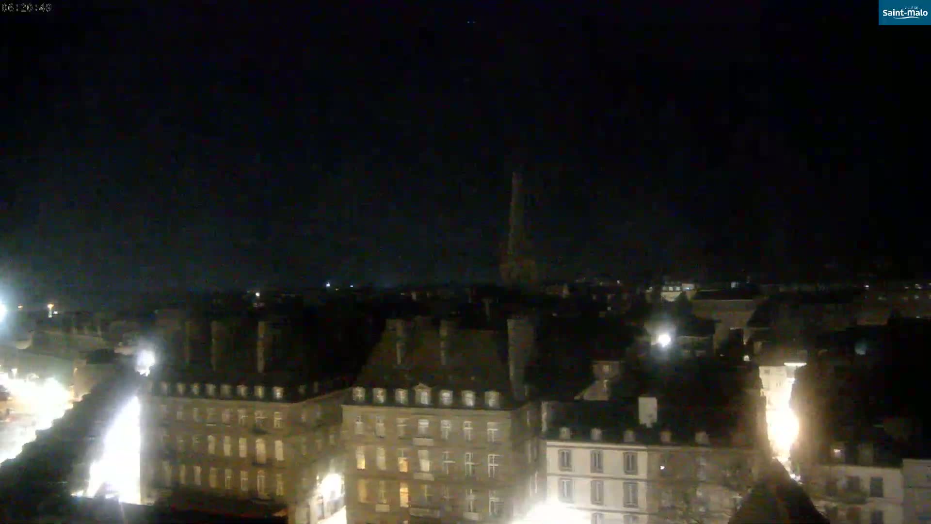 A nighttime, aerial view of a city with many closely-packed buildings, lit with faint streetlights, and a tall spire in the distance under a clear, dark sky.