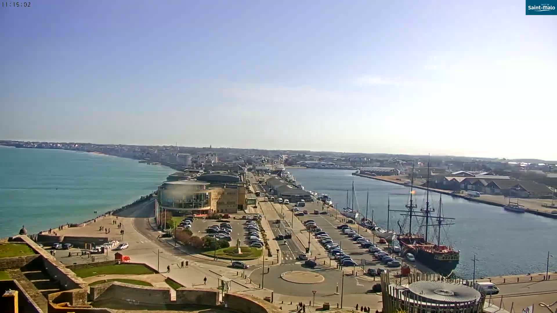 A sunny day overlooking a harbor with a large sailing ship, a waterfront promenade lined with parked cars, and a coastal city extending into the distance.