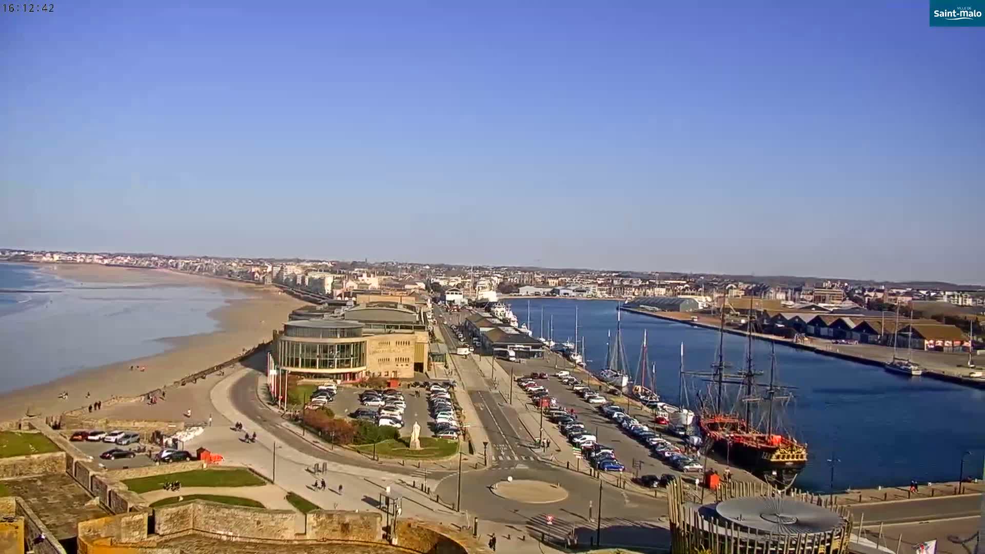 A sunny day reveals a coastal city with a sandy beach, harbor filled with boats, and a large building near the waterfront.