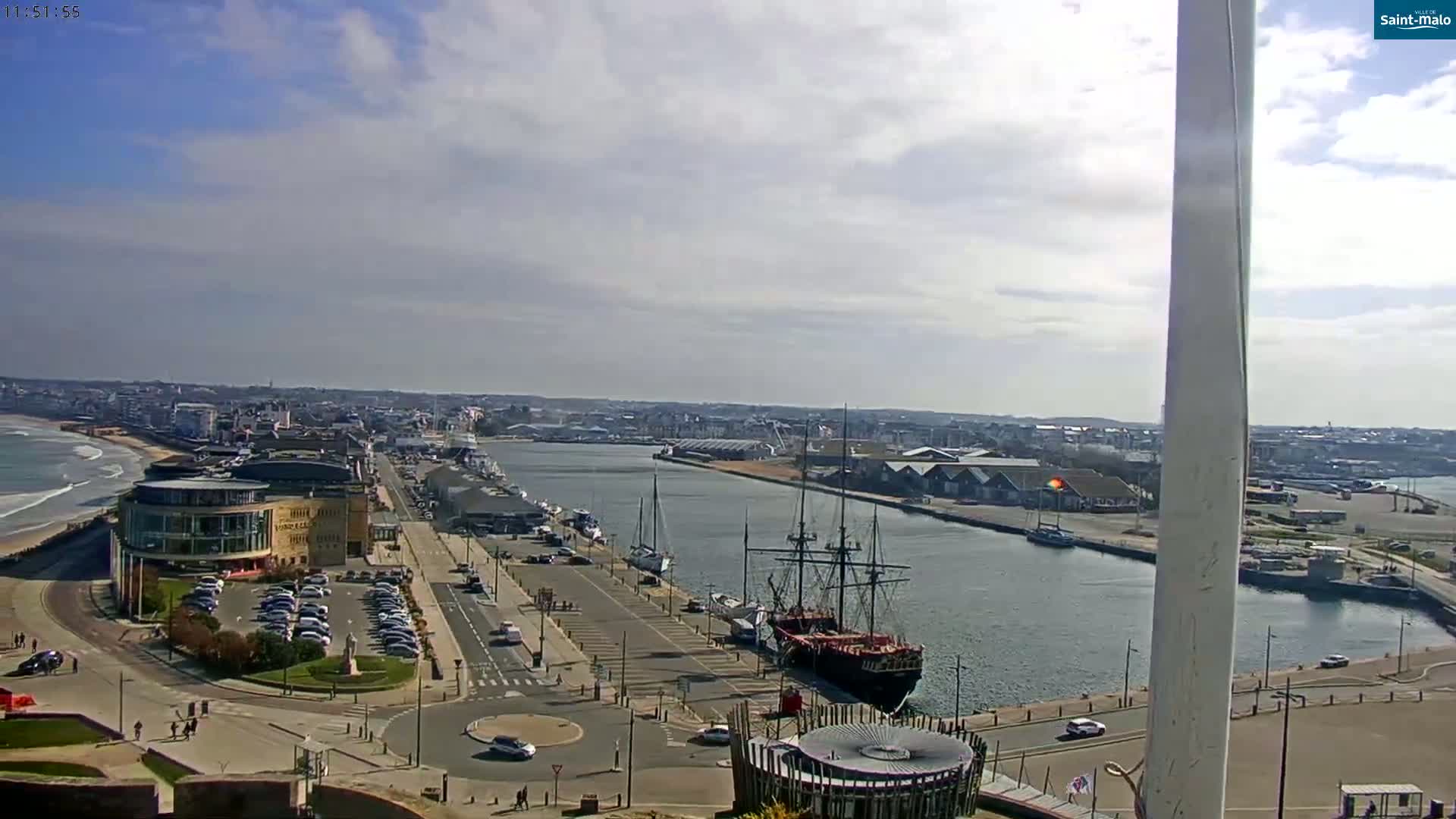 A mostly sunny day overlooks a harbor with a large sailing ship docked near a city with a wide paved area leading to a beach with waves.