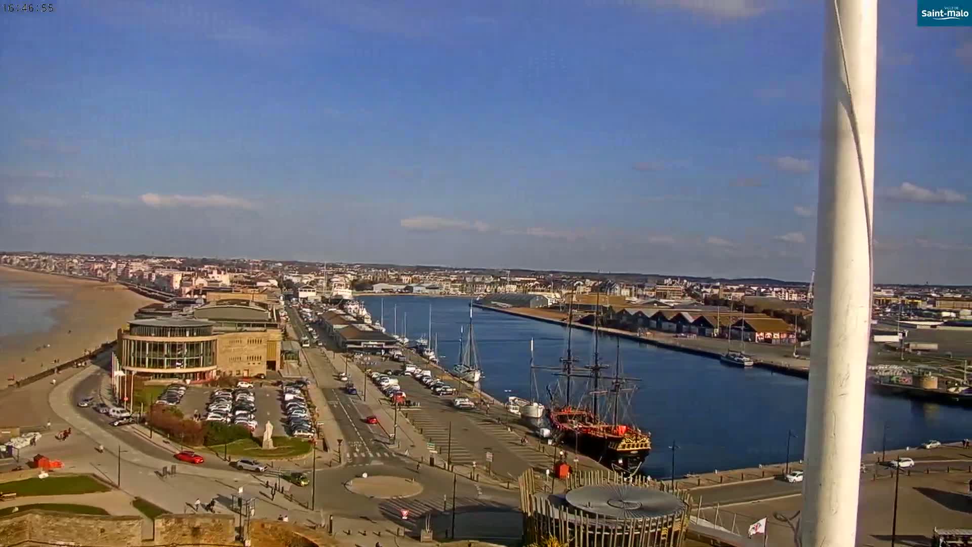 A coastal city with a harbor filled with boats, a sandy beach, and a large building is visible on a partly sunny day.