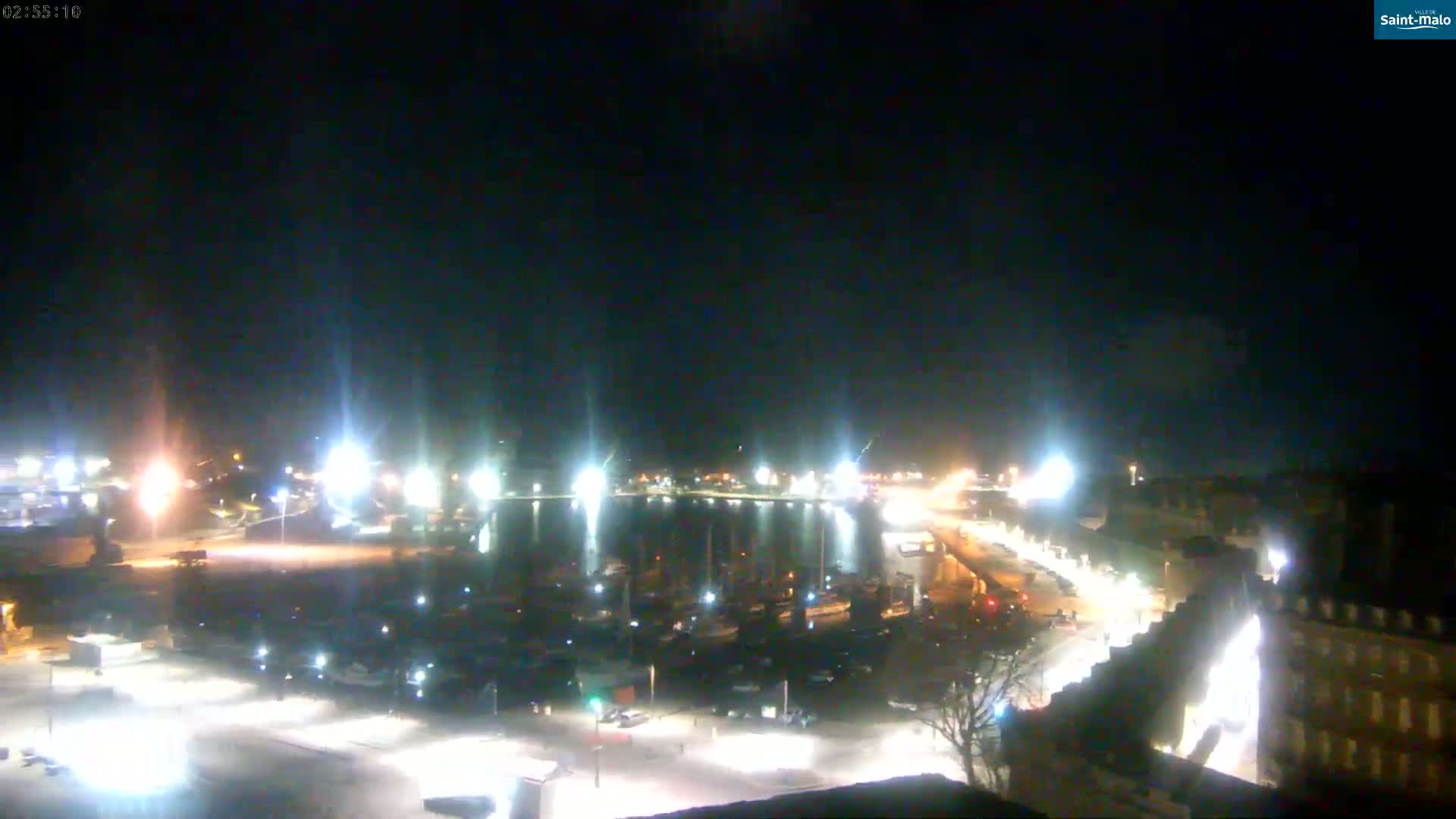 A nighttime, brightly lit harbor filled with boats is seen from an elevated vantage point under a clear sky.